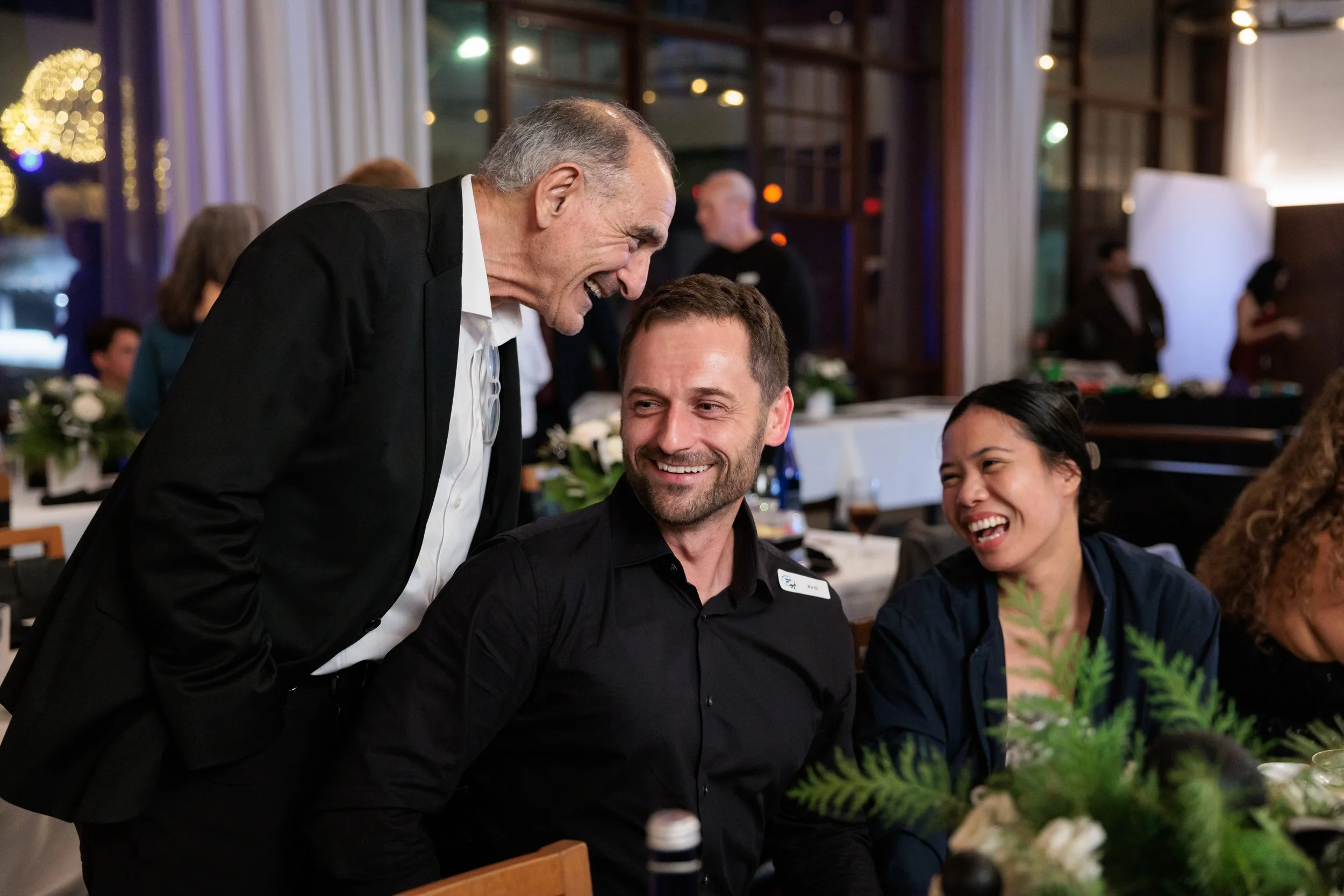 Three people at a social gathering are engaged in conversation and laughing. An older man in a black suit is leaning over to talk to a seated man in a black shirt, both smiling, while a woman with dark hair next to them is also smiling and laughing.