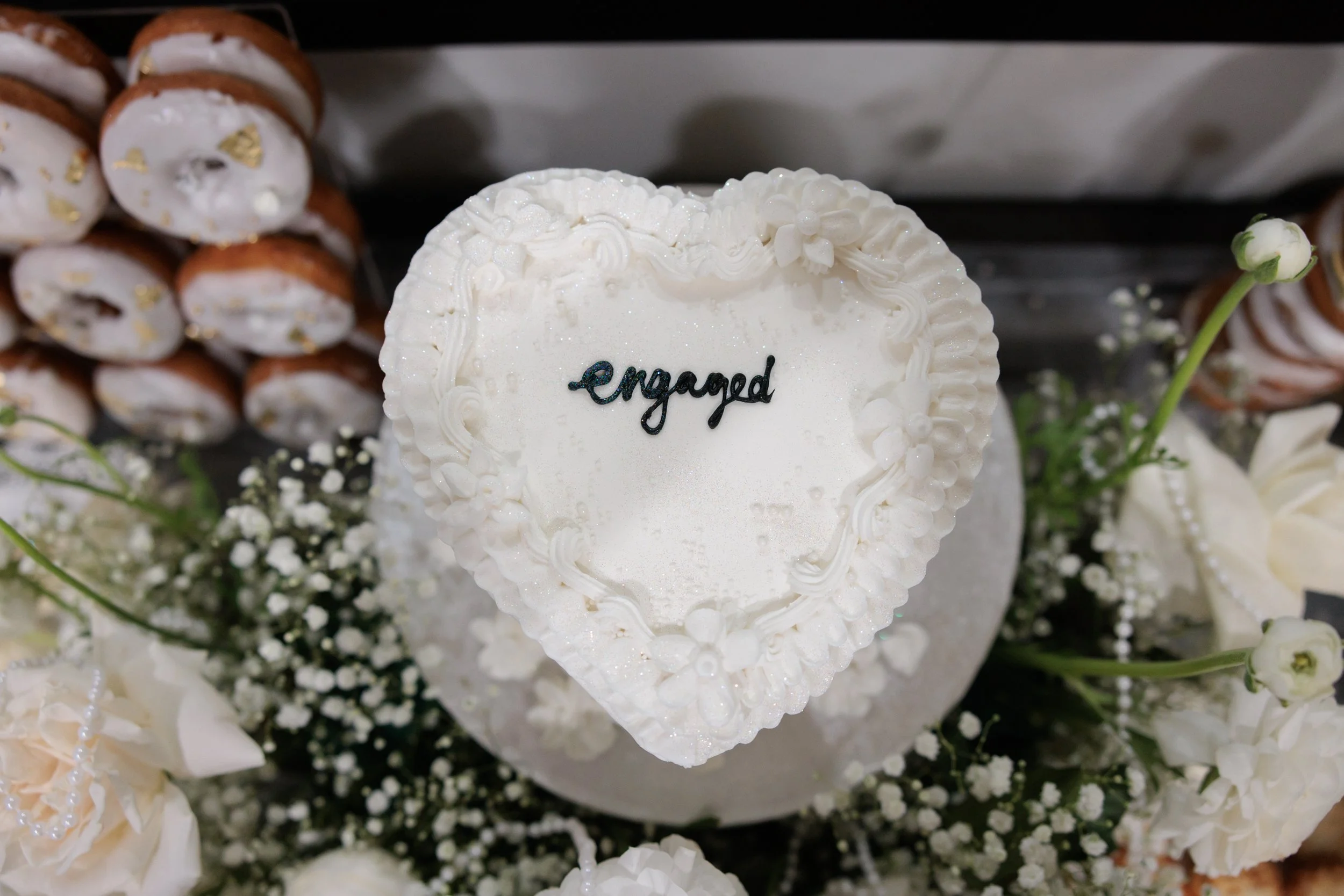Heart-shaped wedding cake with white frosting and decorative flowers, with the word 'engaged' written on top, surrounded by white flowers and bakery items in the background.