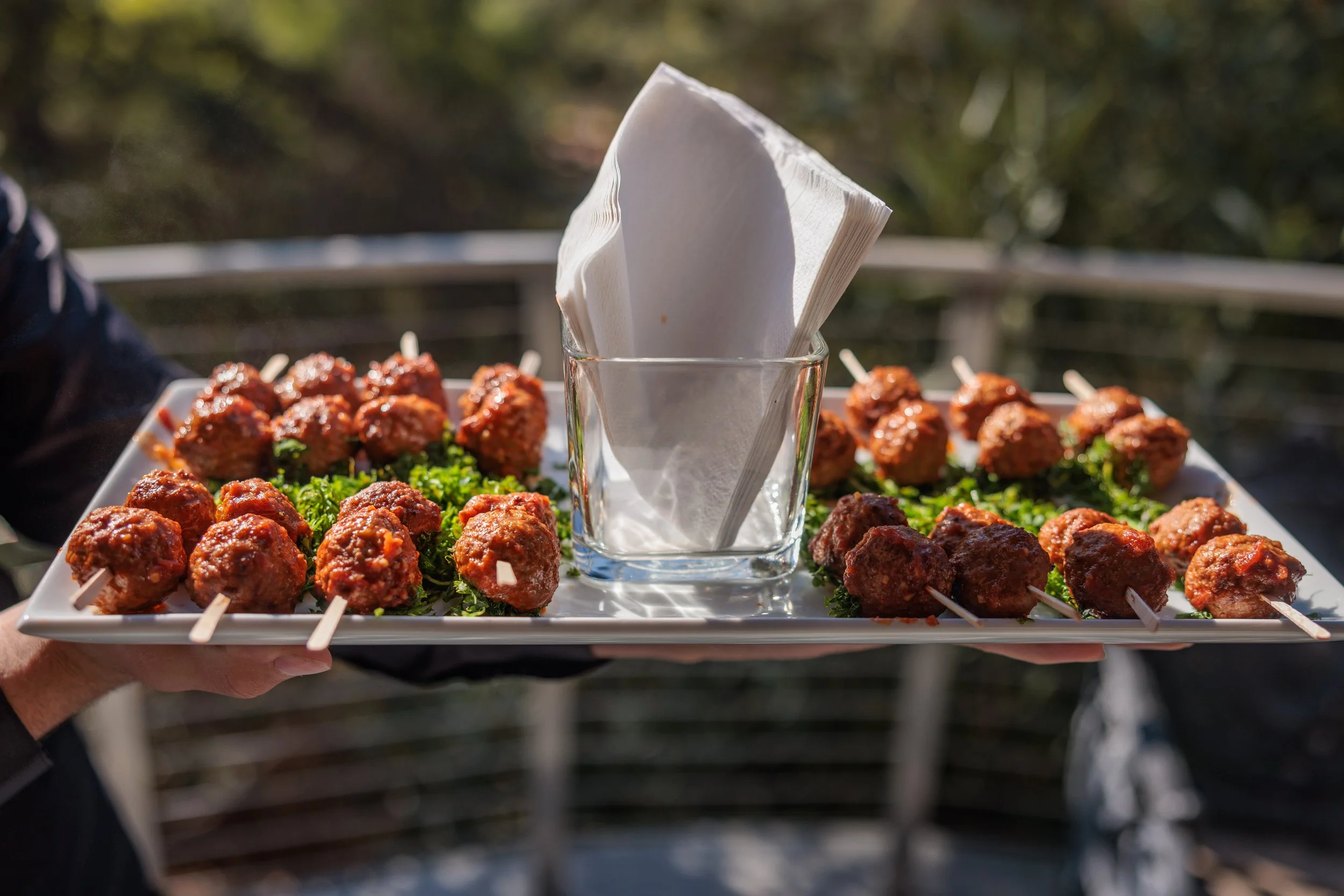 A person holds a tray of meatballs on skewers arranged on a bed of parsley, with a glass containing napkins in the center, outdoors.