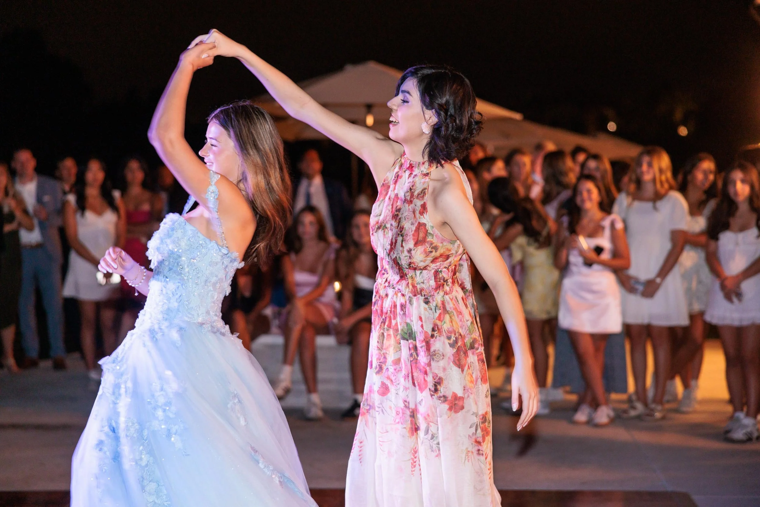 Two women dancing at an outdoor party at night, with a crowd of onlookers in casual and semi-formal attire in the background.