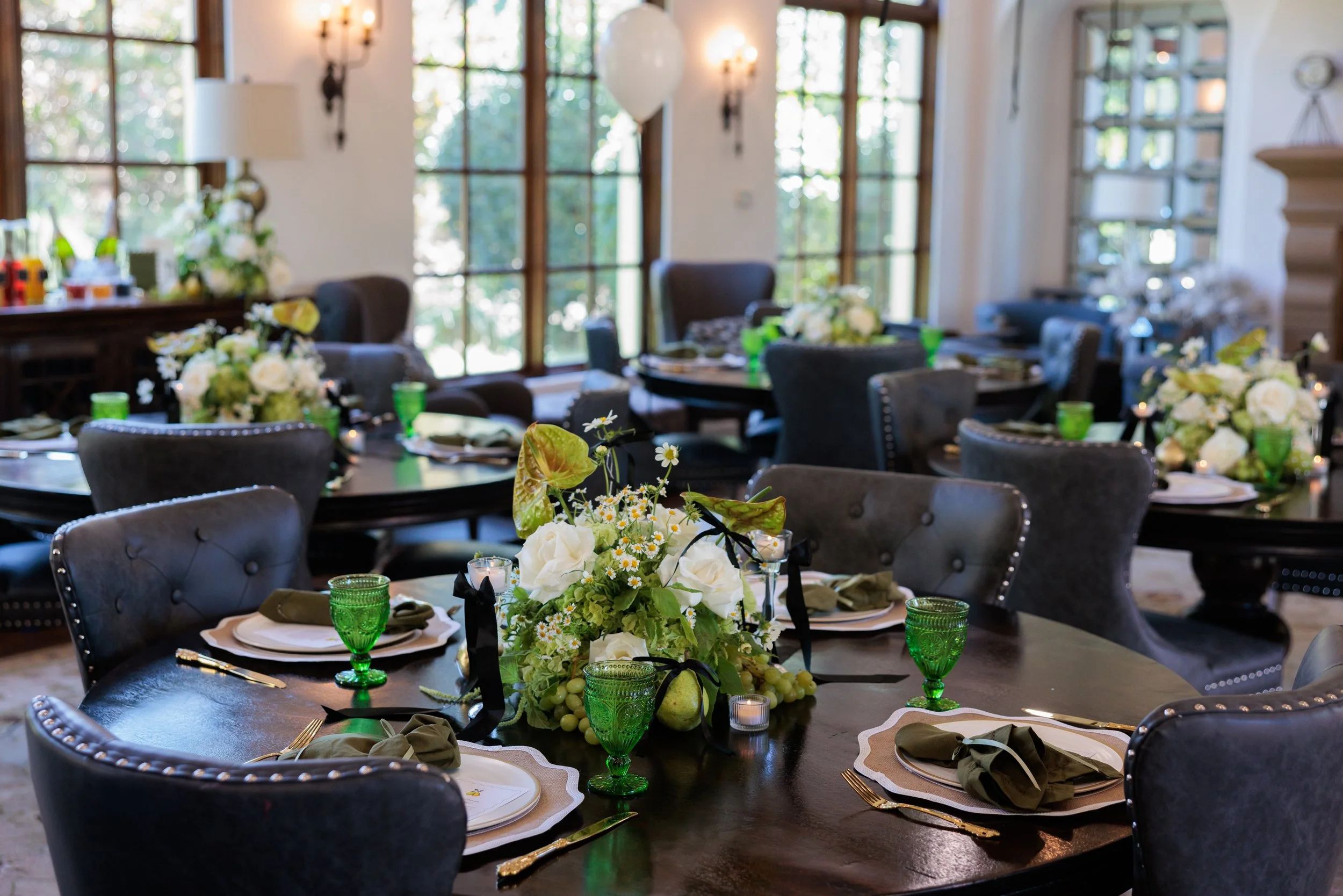 Elegant dining table setup for a wedding or celebration with floral centerpieces, green glassware, and place settings in a bright room with large windows.