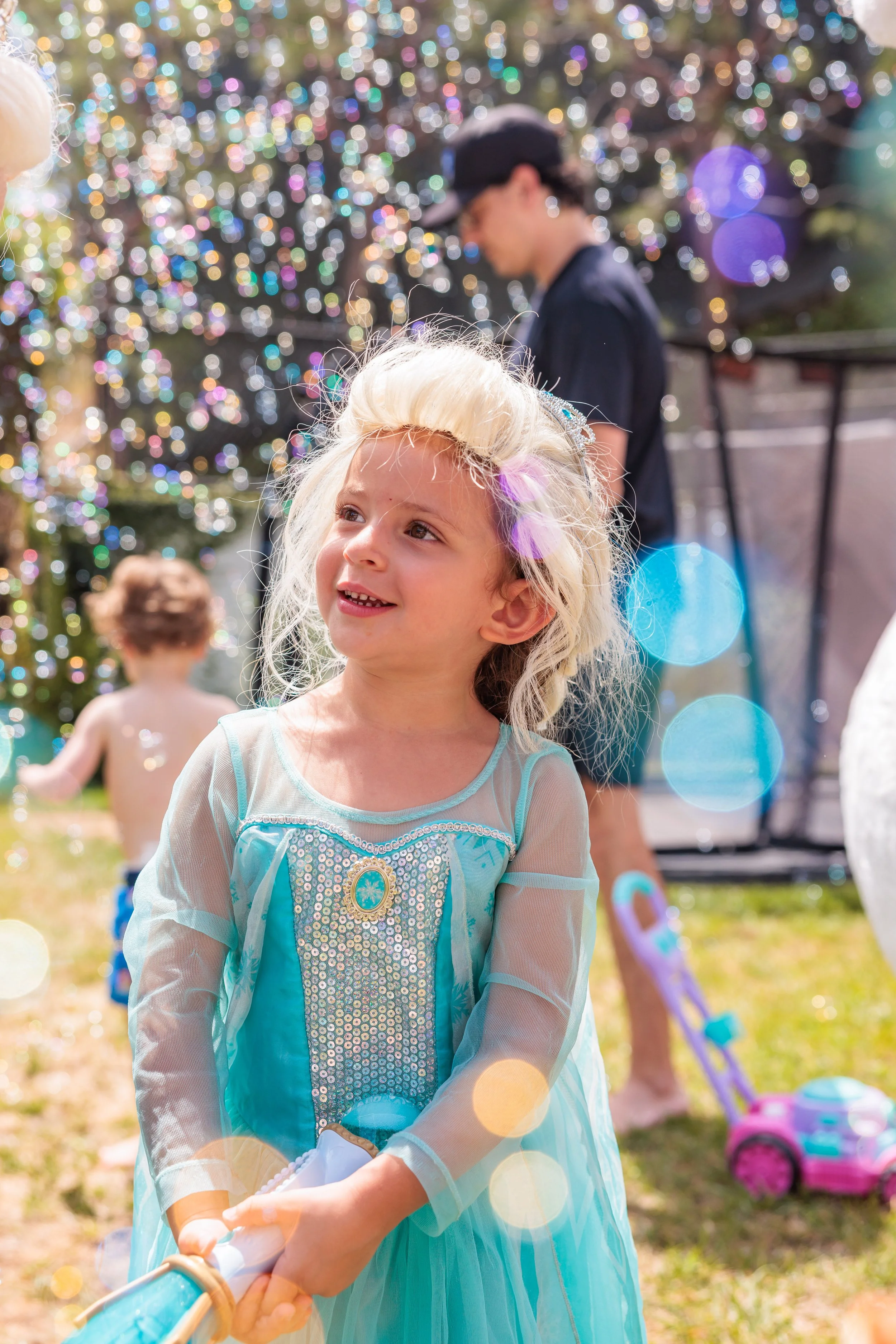 Young girl dressed as Elsa from Frozen smiling outdoors on a sunny day, holding a toy, with other children and a Christmas tree decorated with colorful lights in the background.