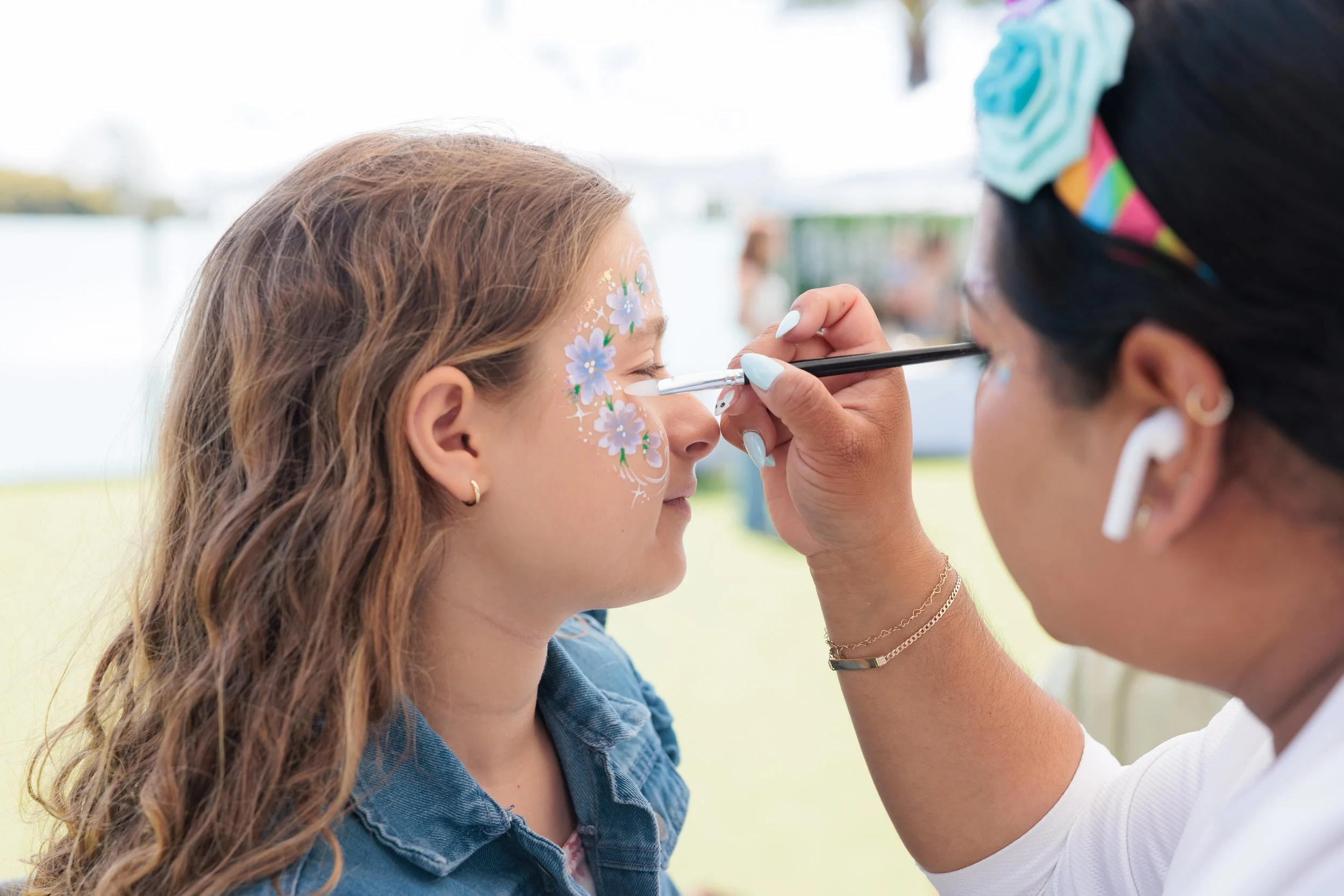 A woman is painting floral face art on a young girl's face outdoors, with the girl sitting still and eyes closed.