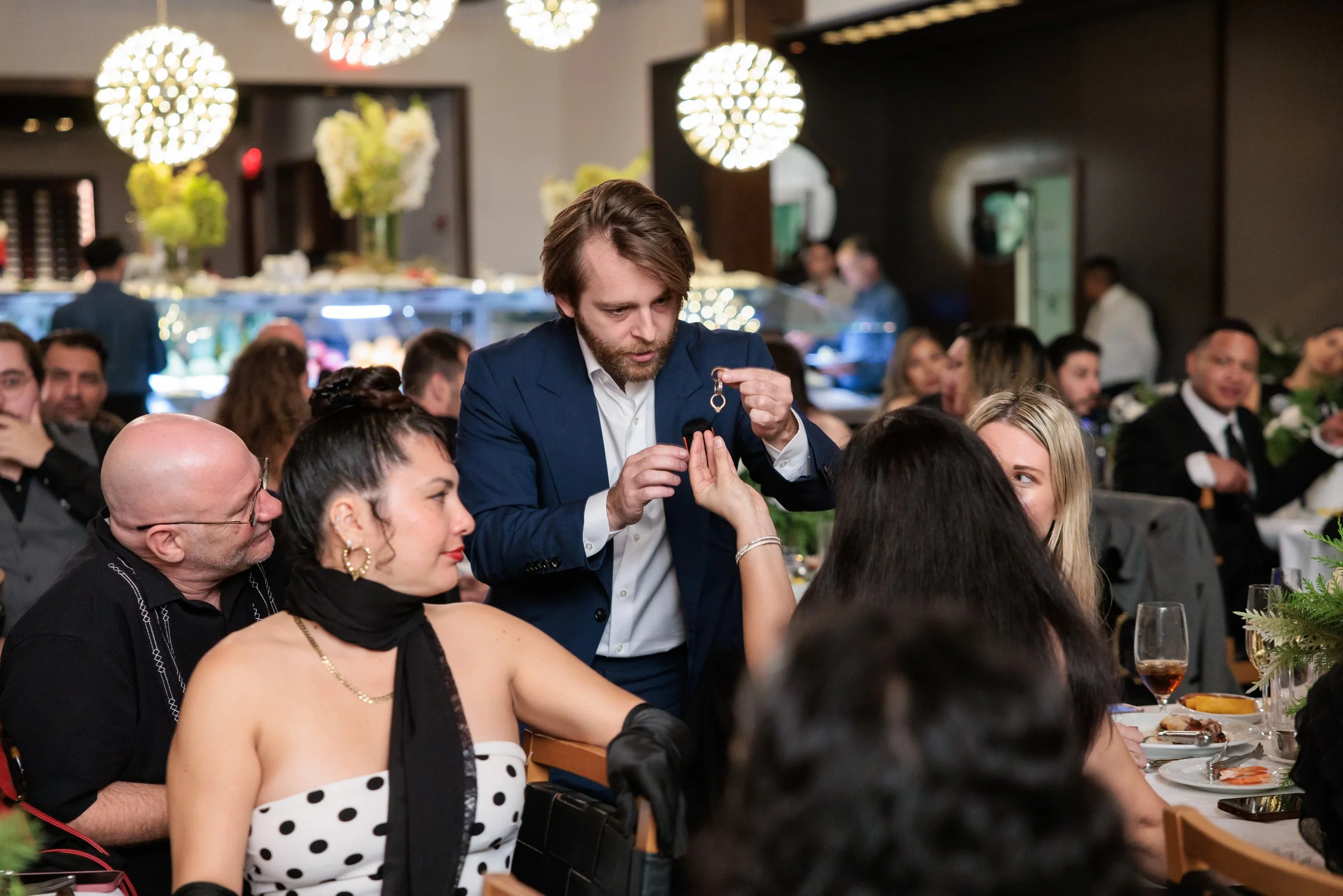 A man in a blue suit is handing a key to a woman in a polka dot dress at a formal gathering or celebration, with other people seated at tables around them.