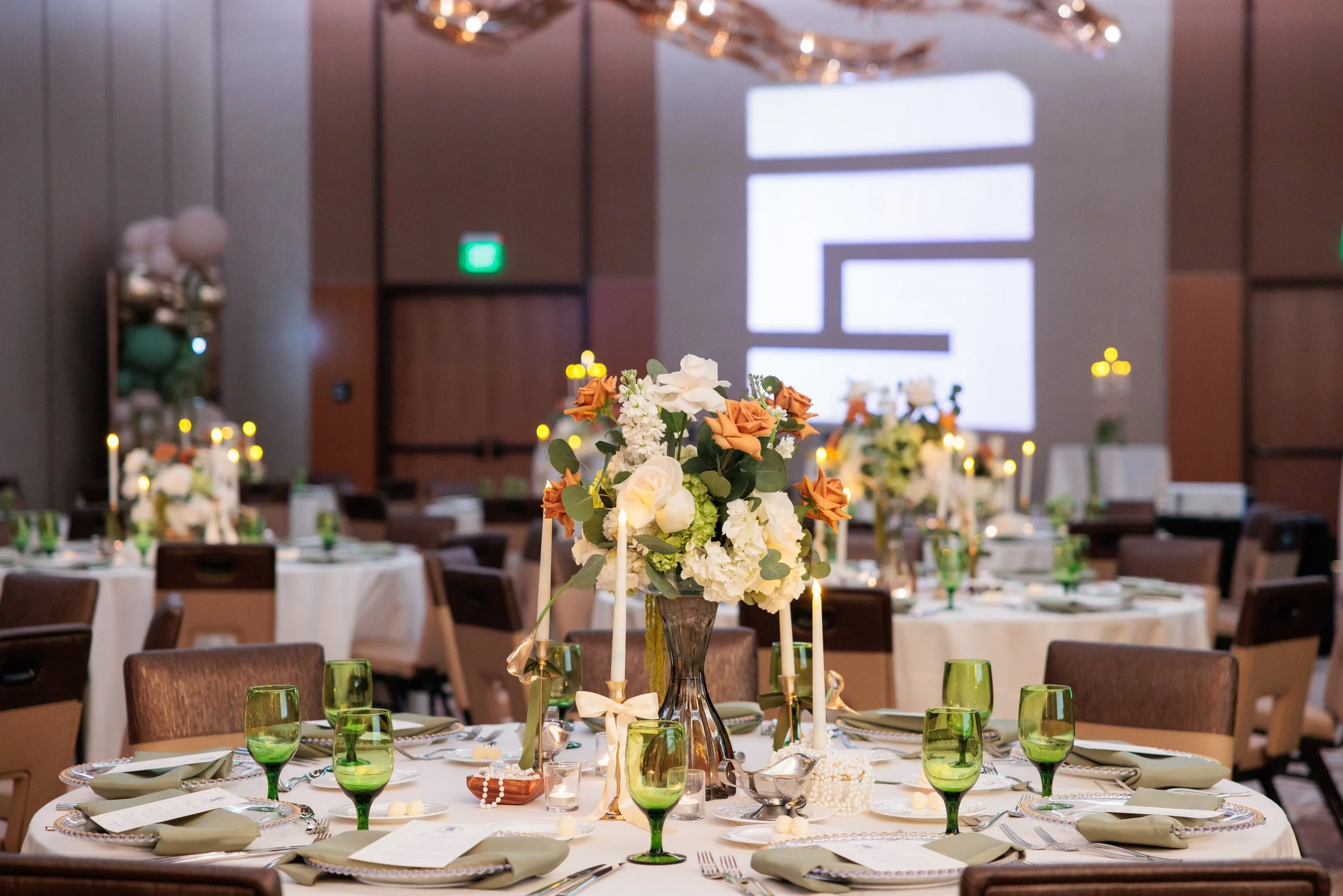 Elegant event banquet table with a centerpiece of white, peach, and orange flowers, surrounded by green glasses and set with plates, utensils, and napkins, in a decorated hall with large digital screen in background.