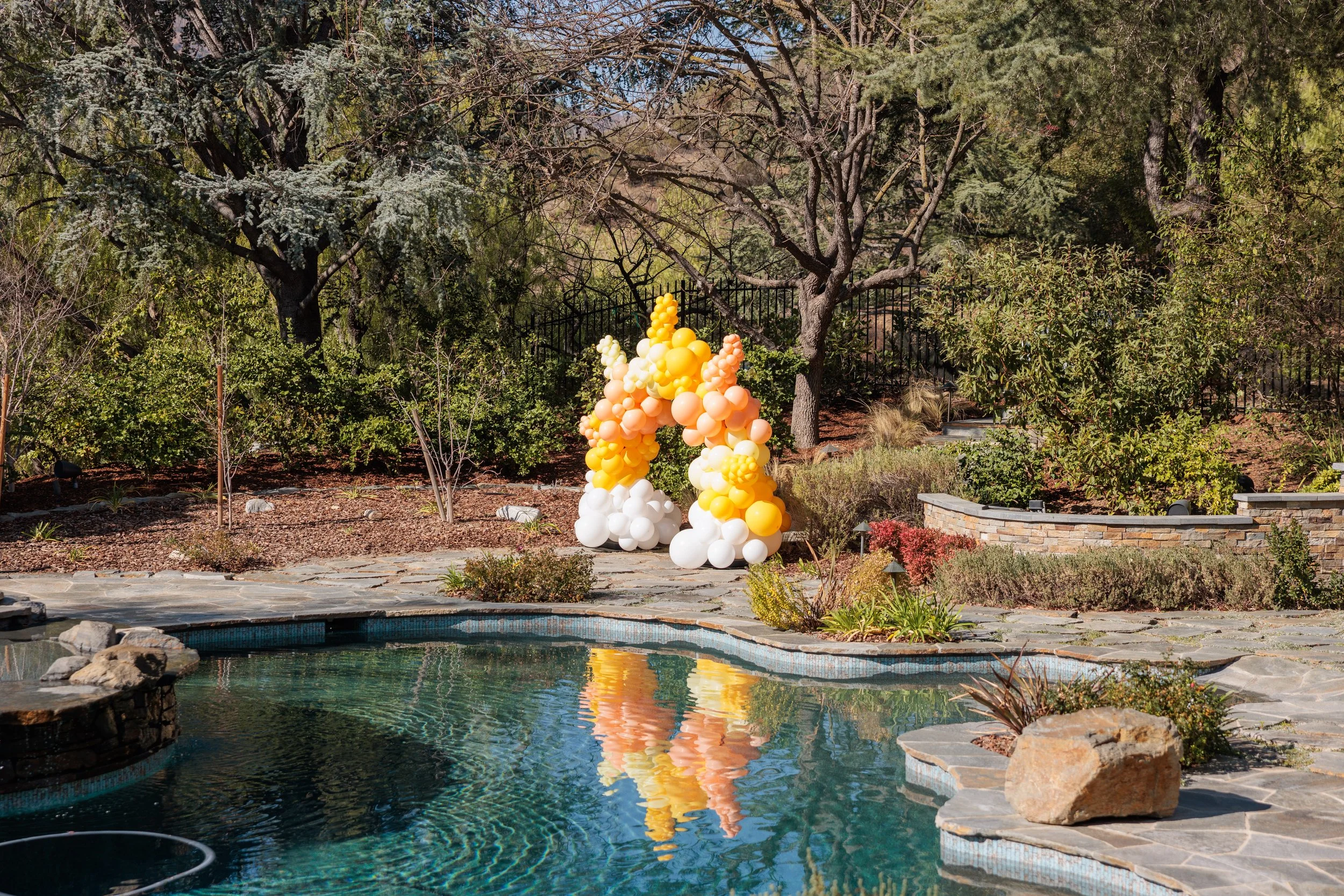Decorative balloon arch with yellow, pink, and white balloons near a backyard garden and swimming pool.