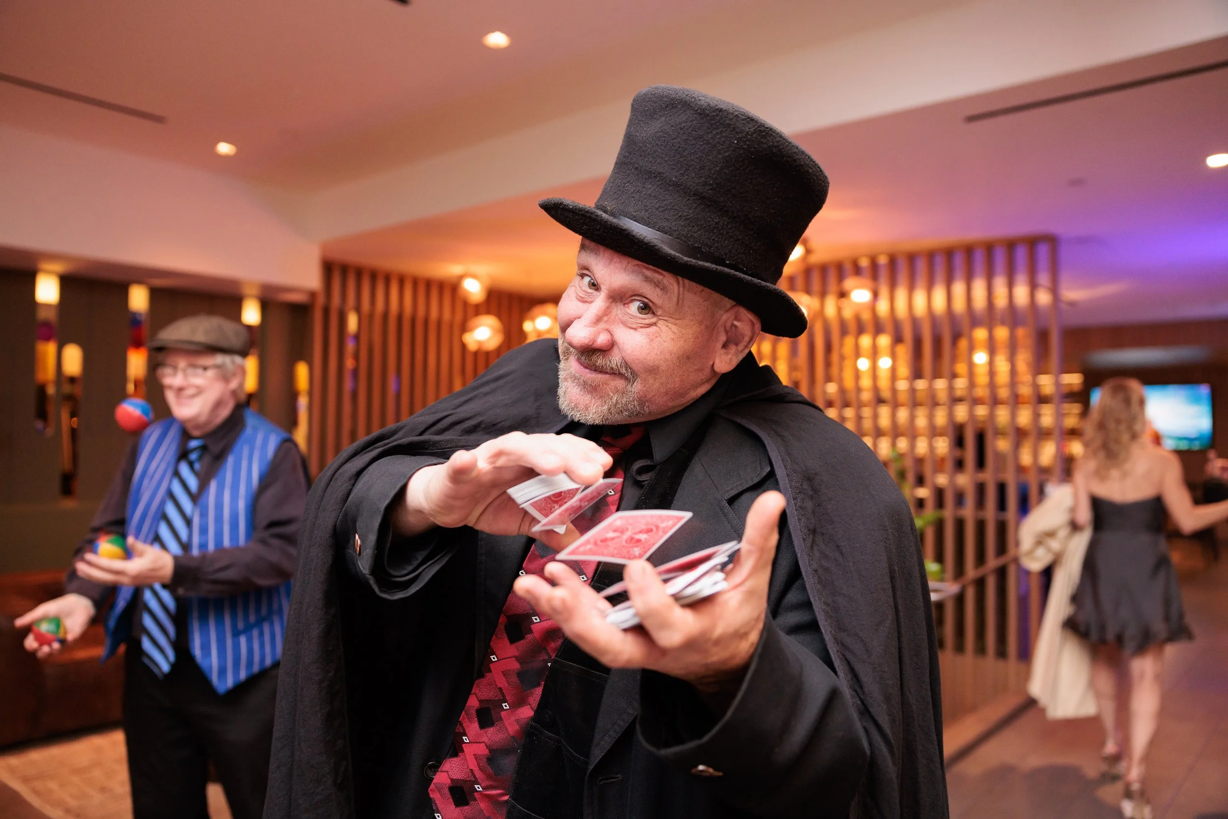 A man wearing a top hat and black coat playfully displaying a deck of cards at a social event, with another man in a blue vest and striped tie juggling colorful balls in the background, in a warmly lit, modern indoor setting.