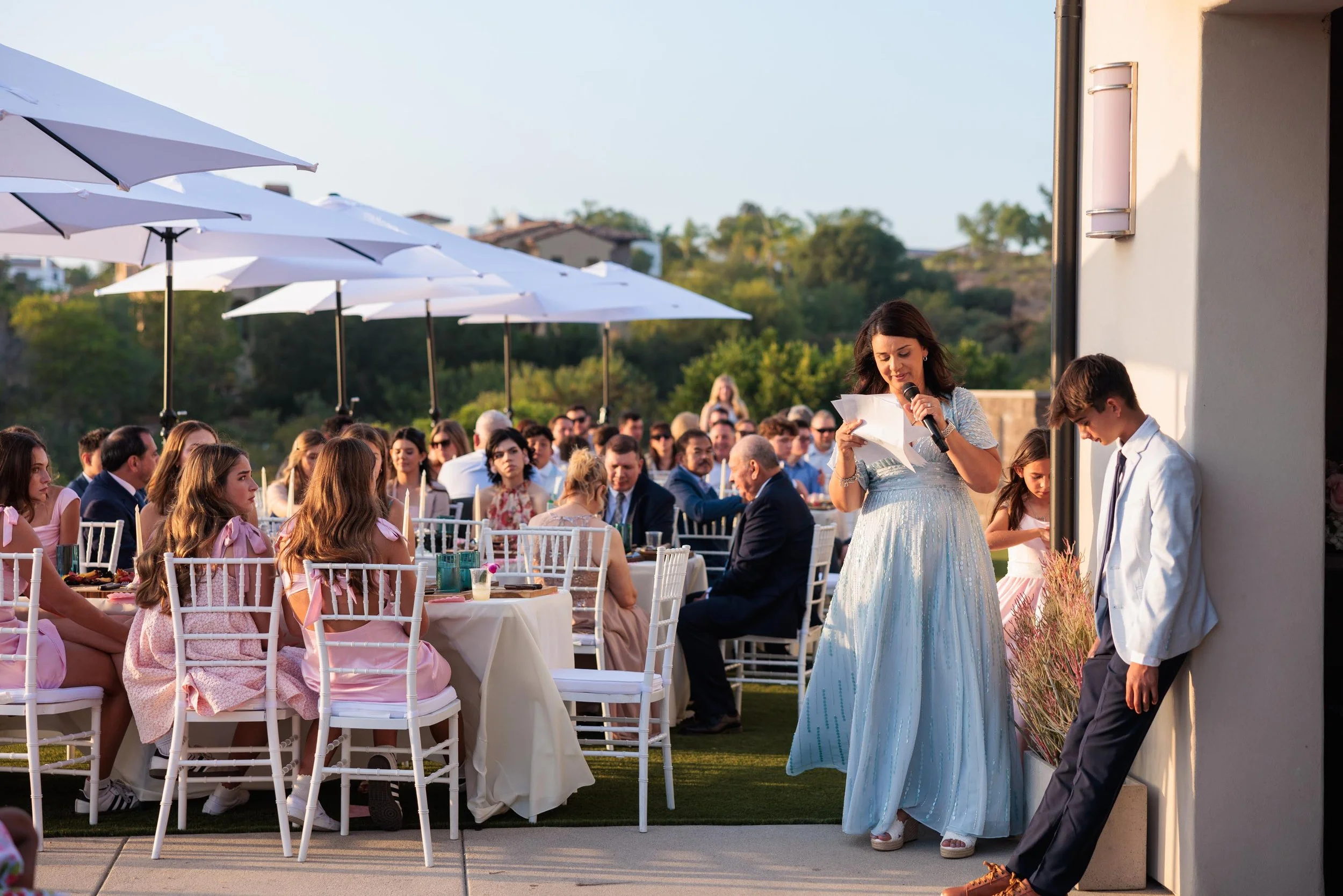 A woman in a silver dress giving a speech at an outdoor wedding reception, standing beside a young man in a white jacket, with guests seated at tables under white umbrellas.