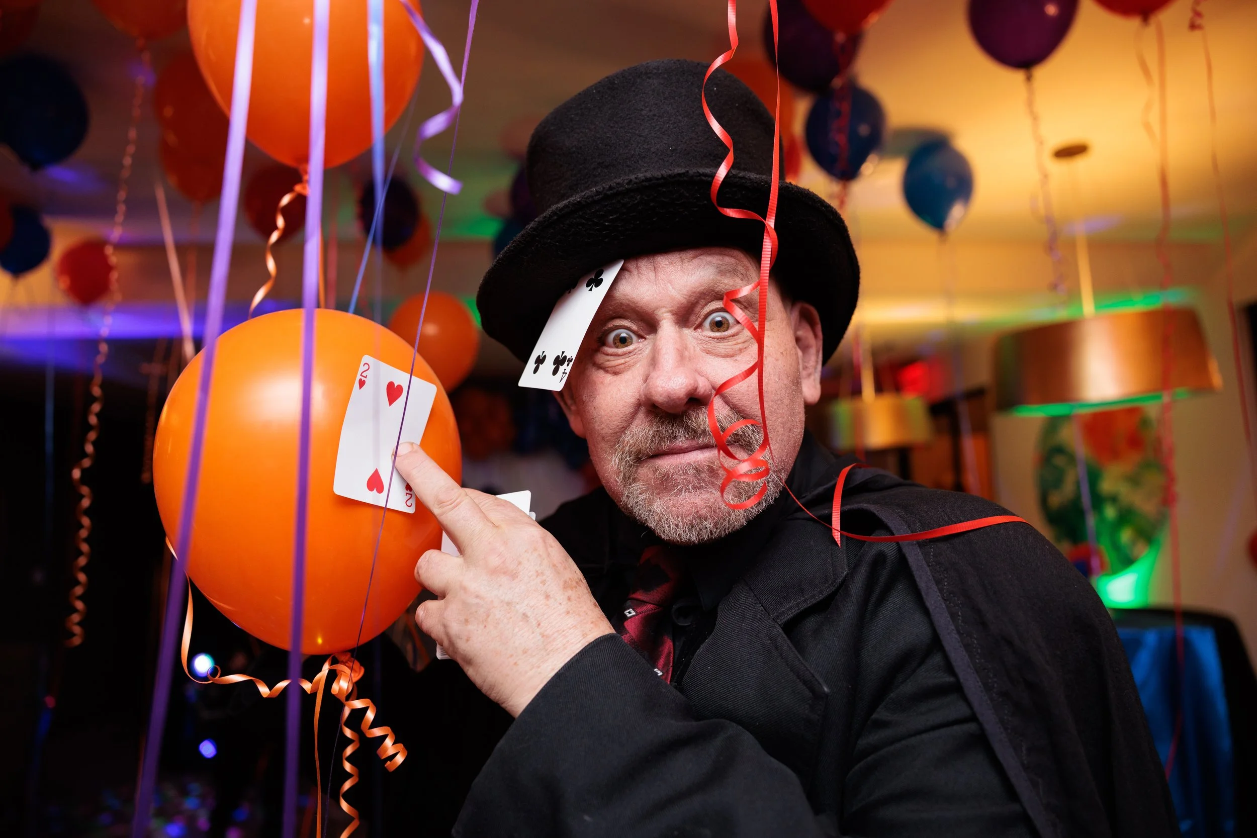 A man dressed as a magician with a black top hat and coat, surrounded by balloons and streamers at a celebration or party, holding playing cards.