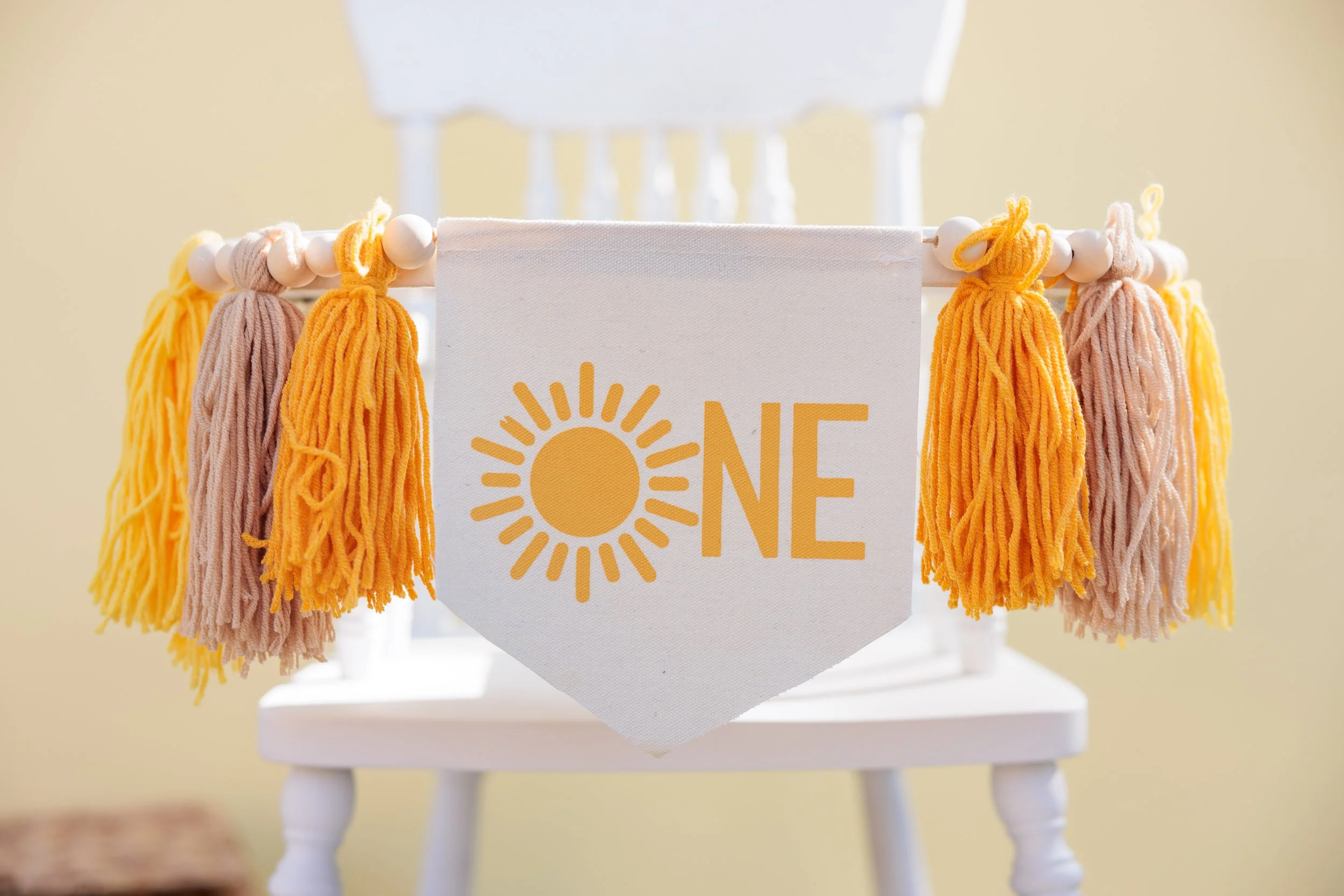 Decorative banner on a white chair with tassels in yellow, pink, and beige, bearing the text 'ONE' with a sun graphic replacing the 'O'.