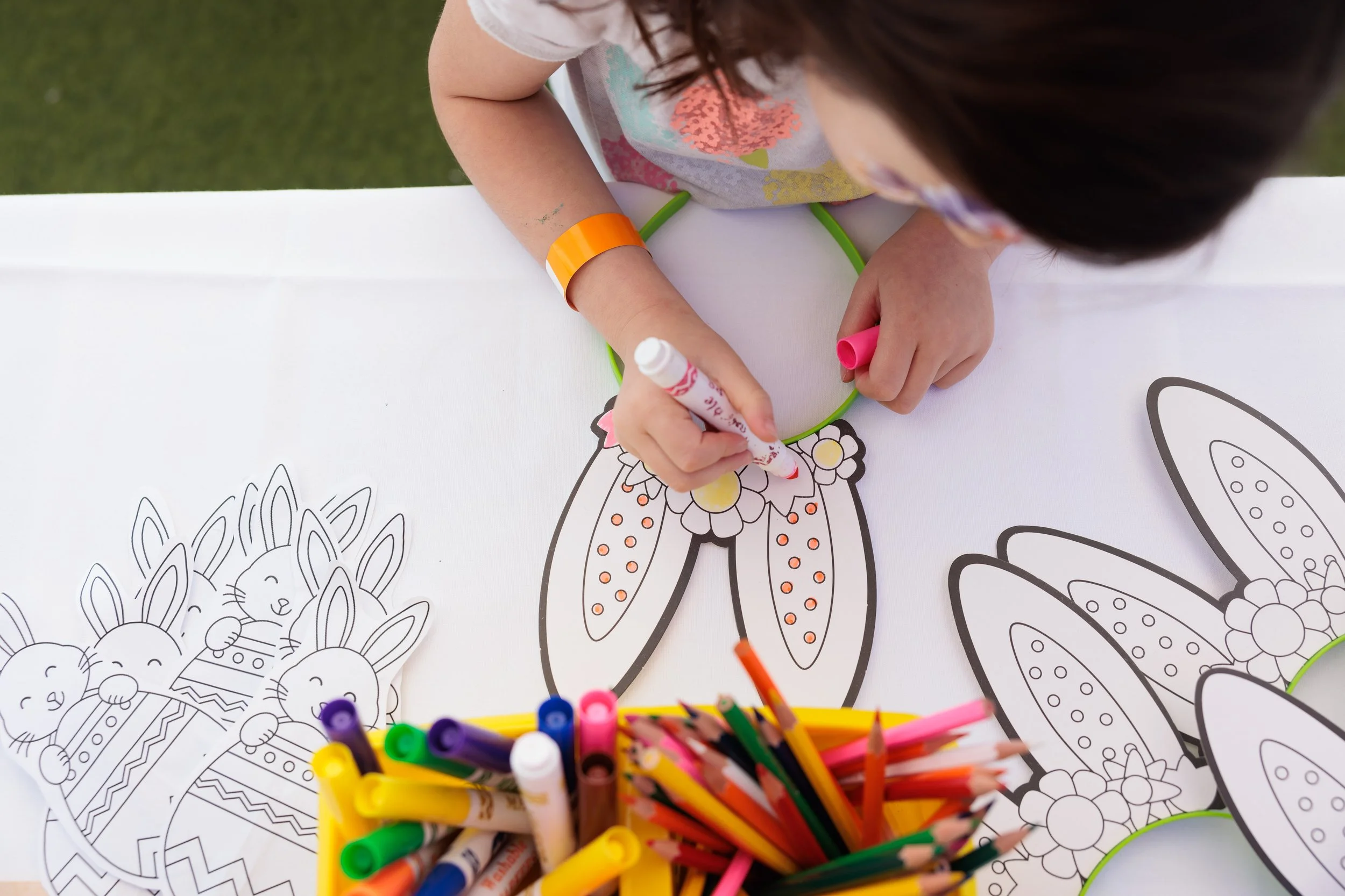 A young girl coloring a butterfly craft at a table with markers and coloring pages of bunnies and other butterfly designs.