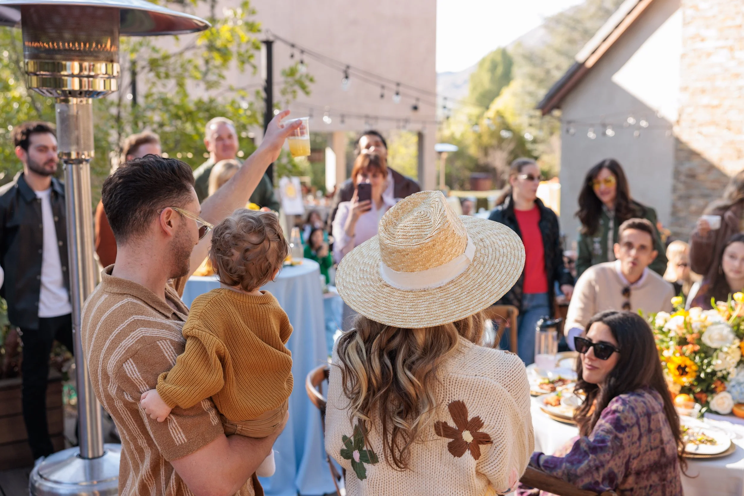 People gathered outdoors at a celebration, including a man holding a child and raising a glass, surrounded by friends and family, with festive decorations and flowers on the table.