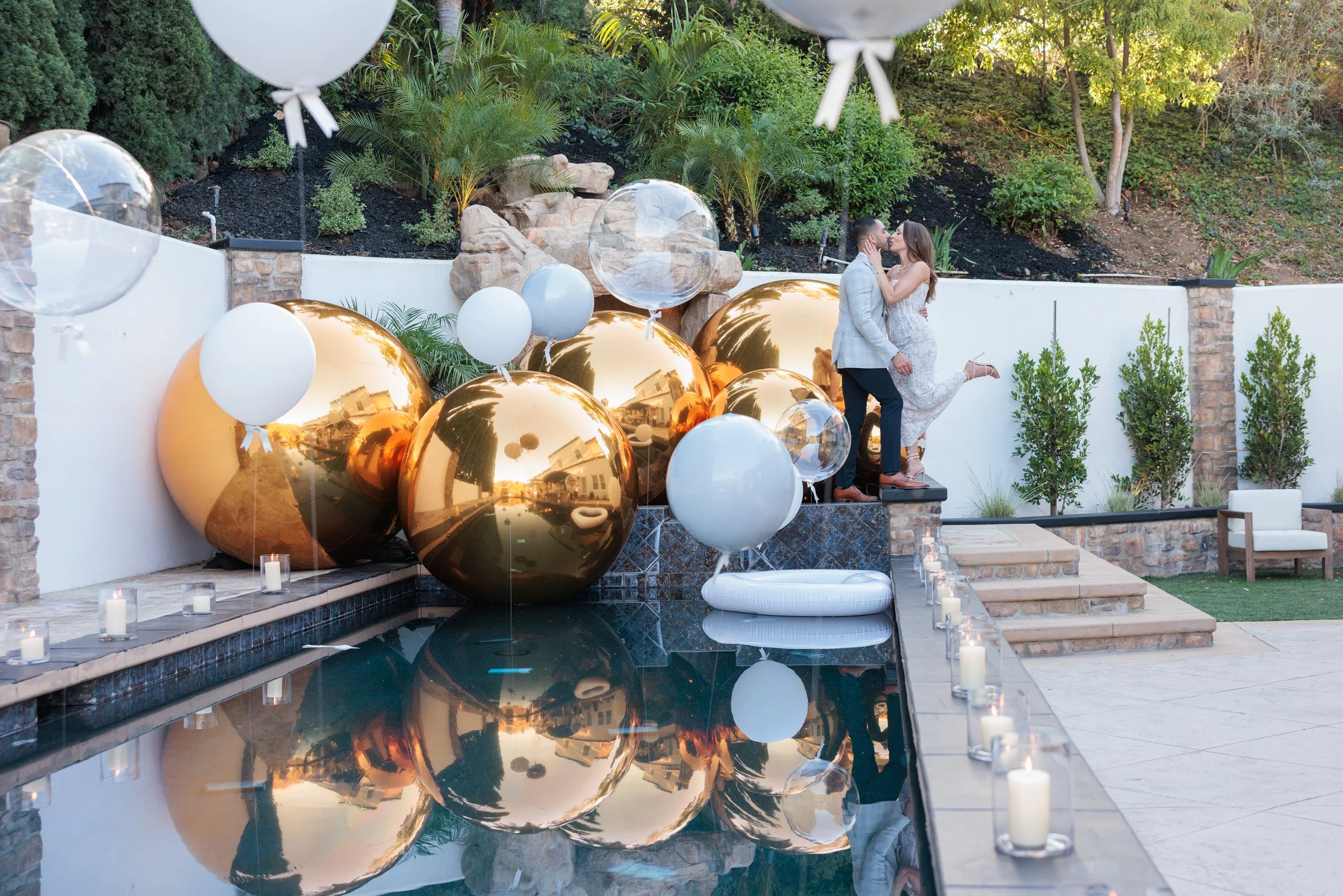 A couple kissing on a ledge before a decorative pool filled with large gold and clear balloons, with candles along the edge and lush greenery in the background.