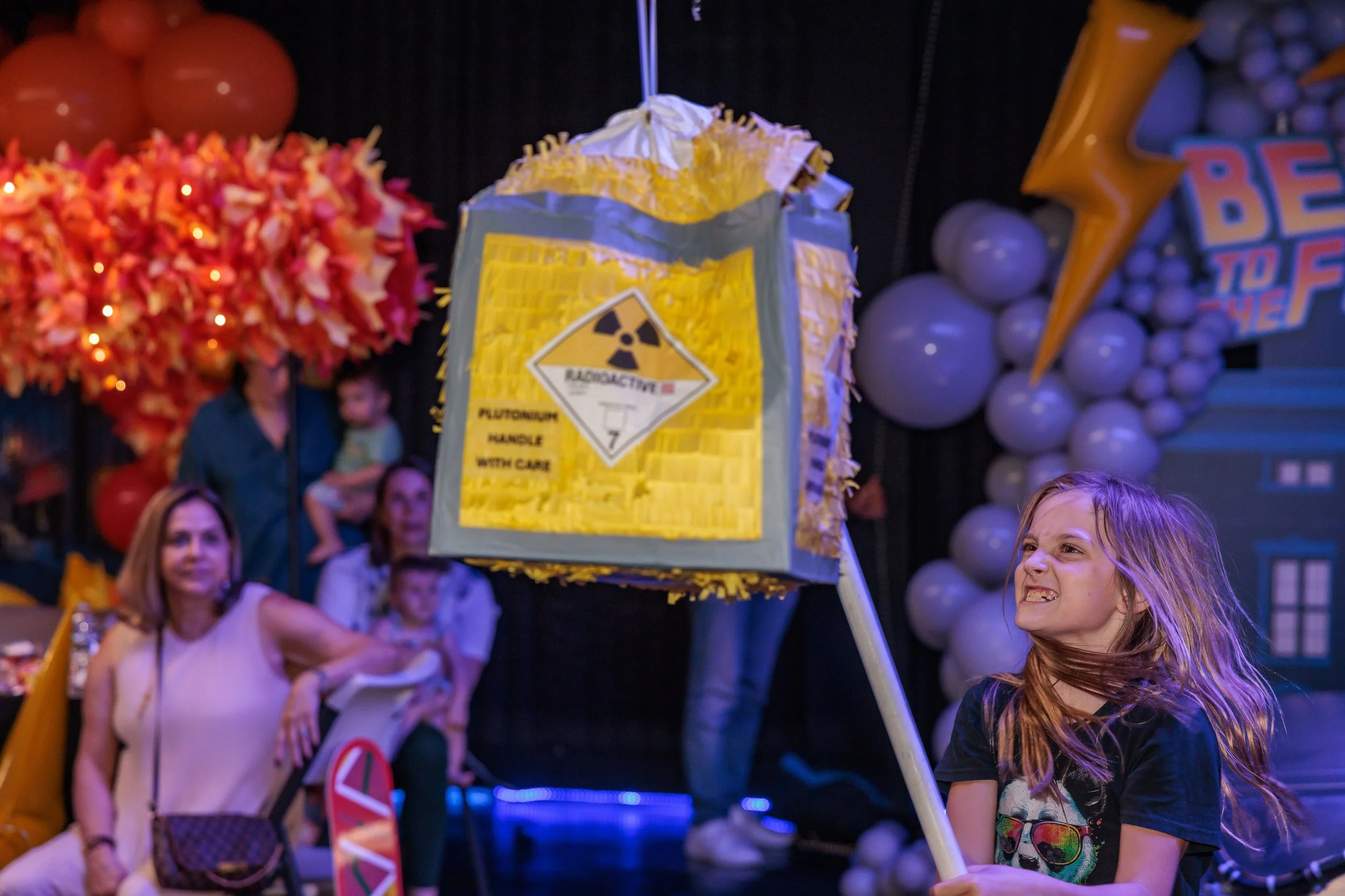 A young girl with long hair is holding a large pollen and radiation hazard sign on a stick during a play or event. There are adults and children in the background, with colorful balloons and decorations, indicating a festive atmosphere.