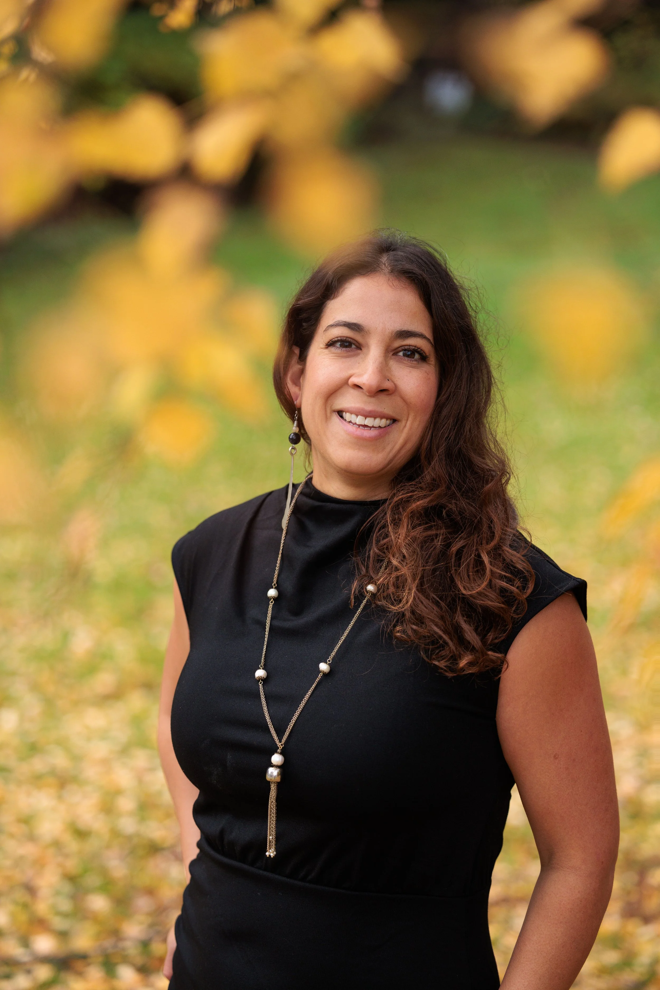 A woman with long dark wavy hair smiling outdoors in front of yellow autumn leaves, wearing a black sleeveless top and a long pearl necklace.
