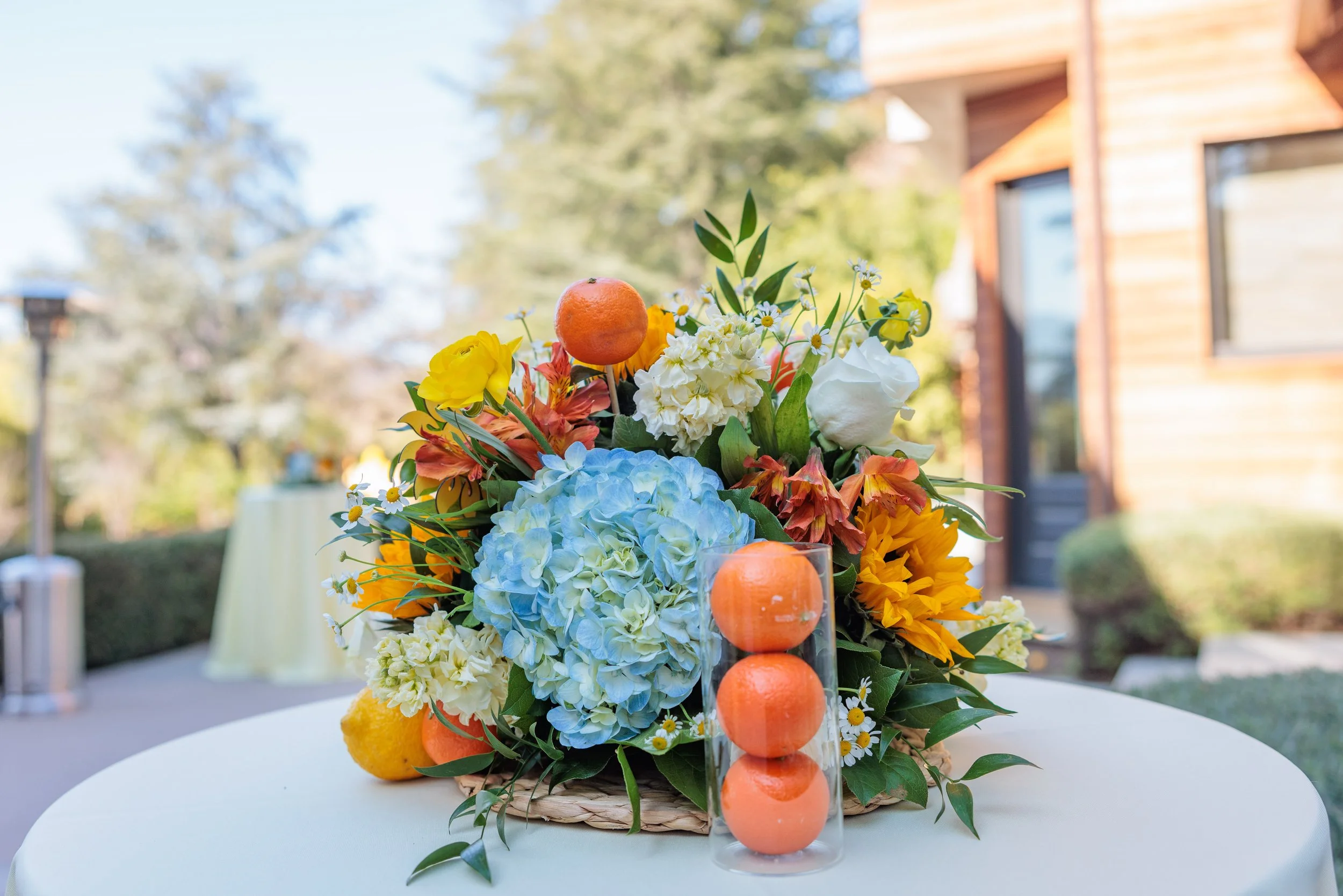 A colorful flower arrangement with blue hydrangeas, yellow sunflowers, white roses, and orange citrus fruits, set on a white table outdoors near a wooden building.