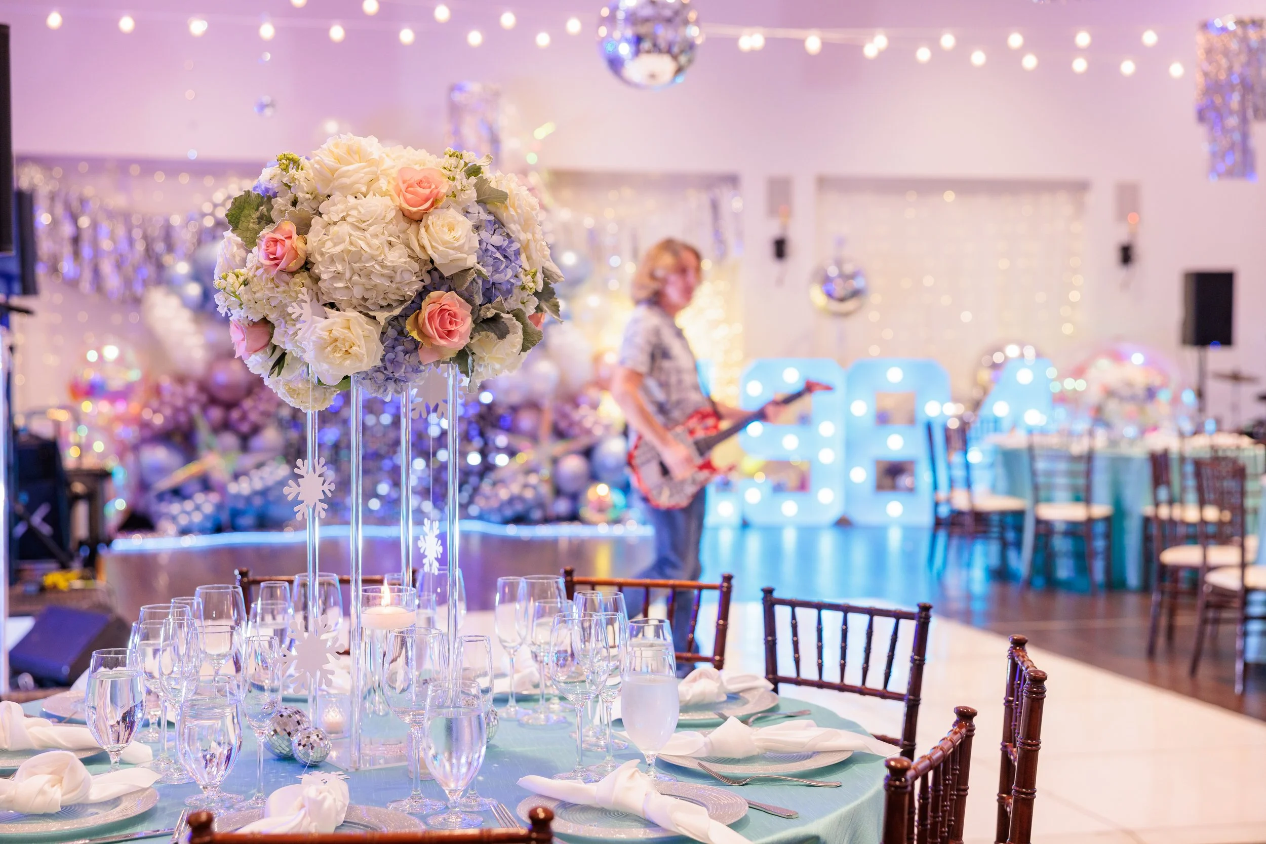 Decorated event space with a table featuring a large floral centerpiece, set with glassware and napkins, and a woman in the background with a guitar, illuminated letters, and Christmas-themed decorations.