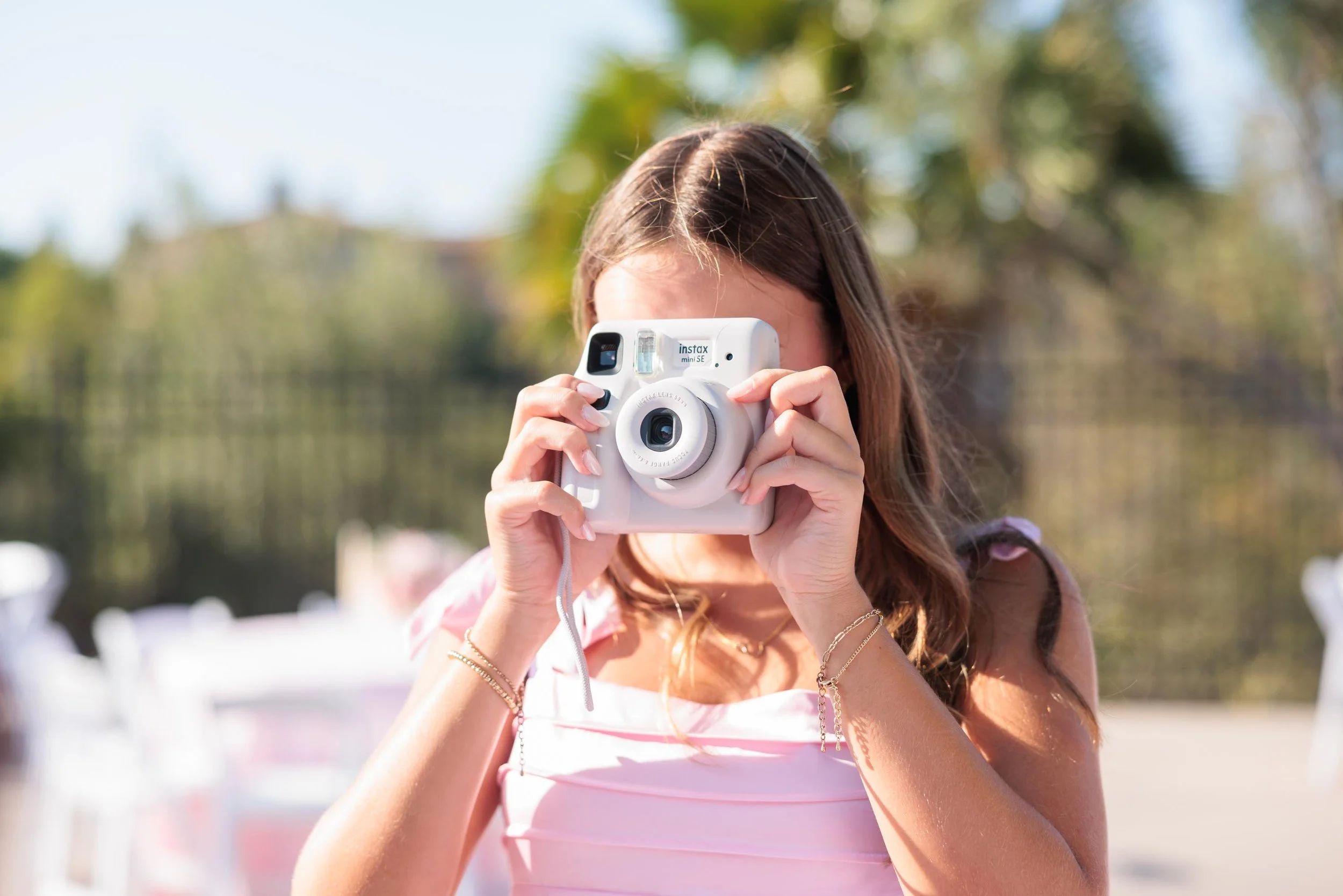 A woman holding a white instant camera and taking a picture outdoors on a sunny day.