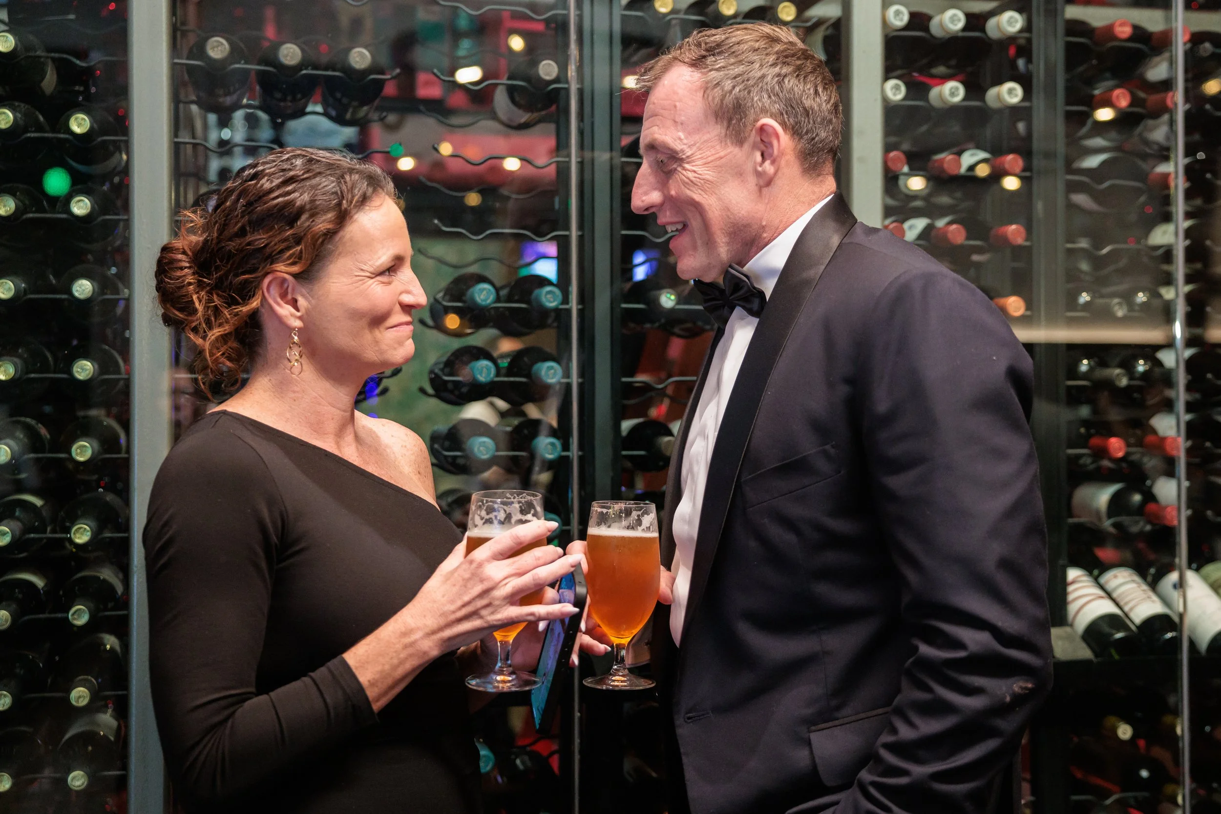 A man and woman in formal attire sharing a toast of beer glasses in front of a wine rack.