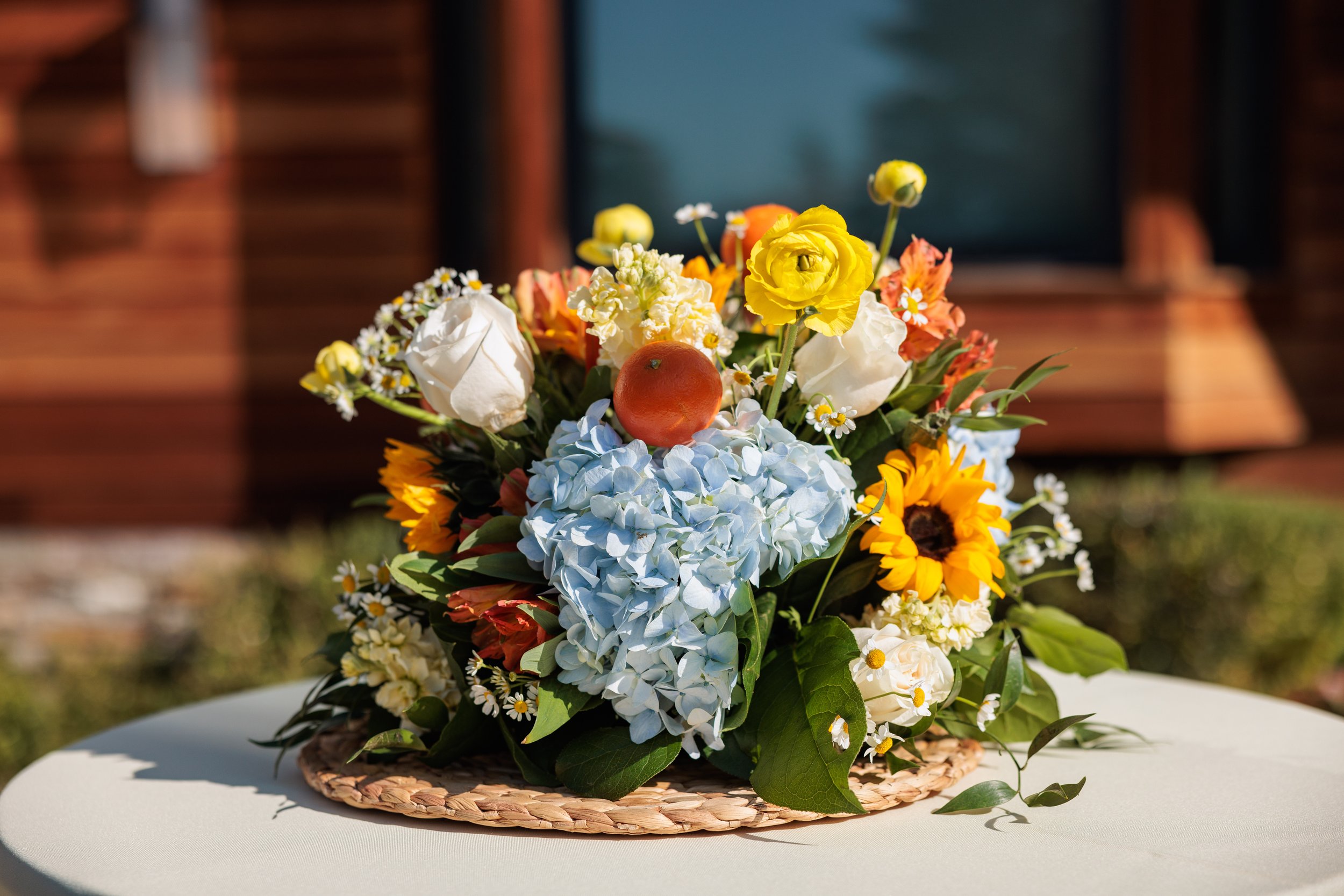 A colorful flower arrangement with hydrangeas, sunflowers, roses, and other flowers and greenery on a white surface.