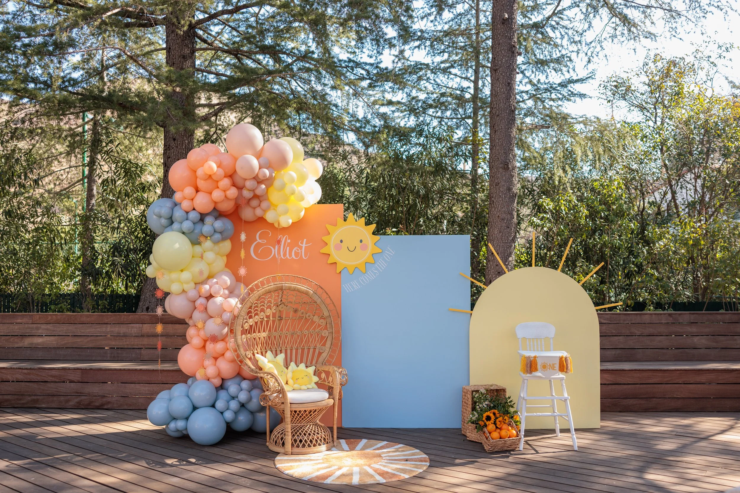Decorative outdoor setup for a children's celebration with pastel balloons, a wicker chair with plush toys, a sun cut-out, and a white chair with a 1st birthday banner, against a background of trees and wooden deck.