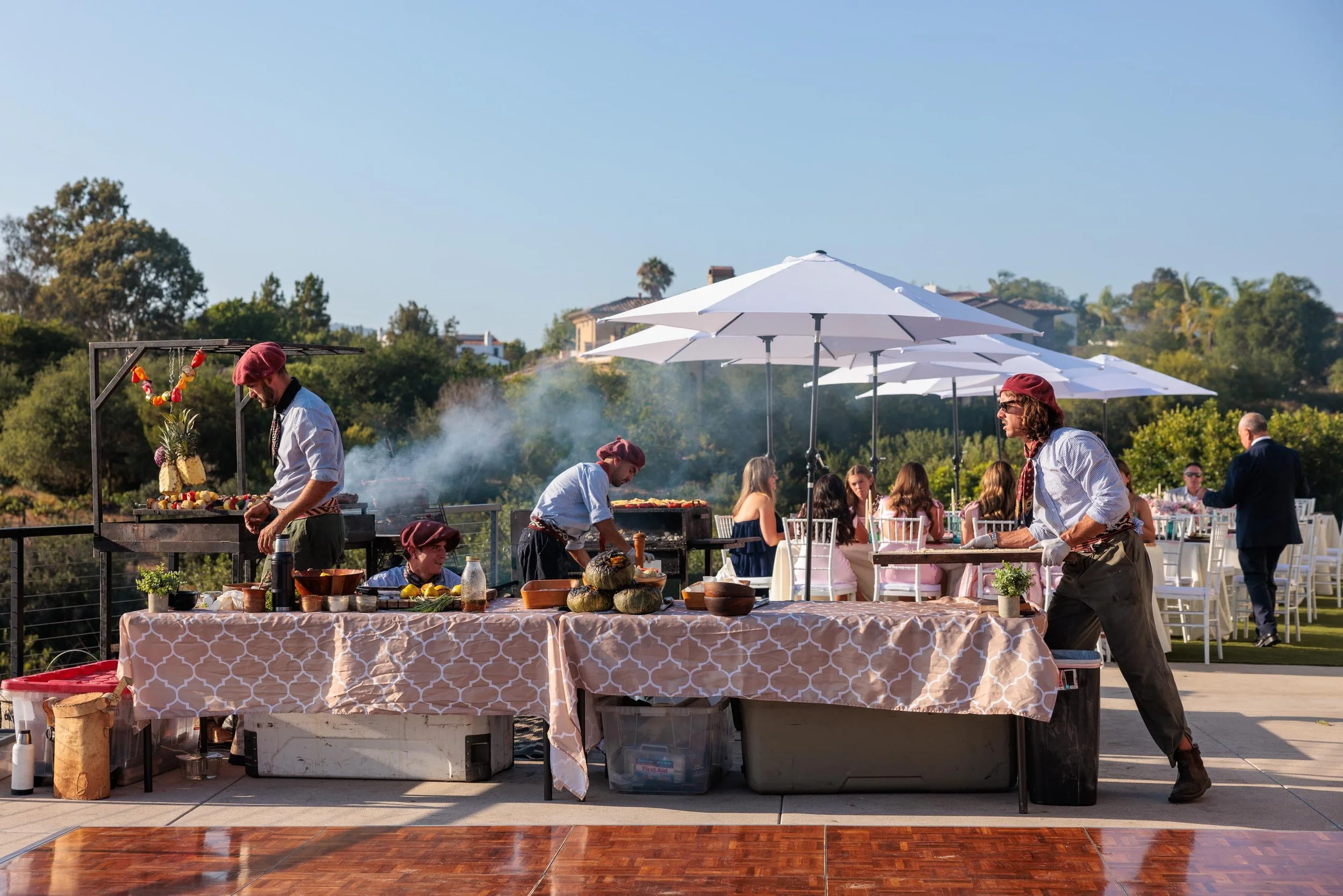 Outdoor catering event with chefs preparing food and guests seated under umbrellas in a scenic setting.