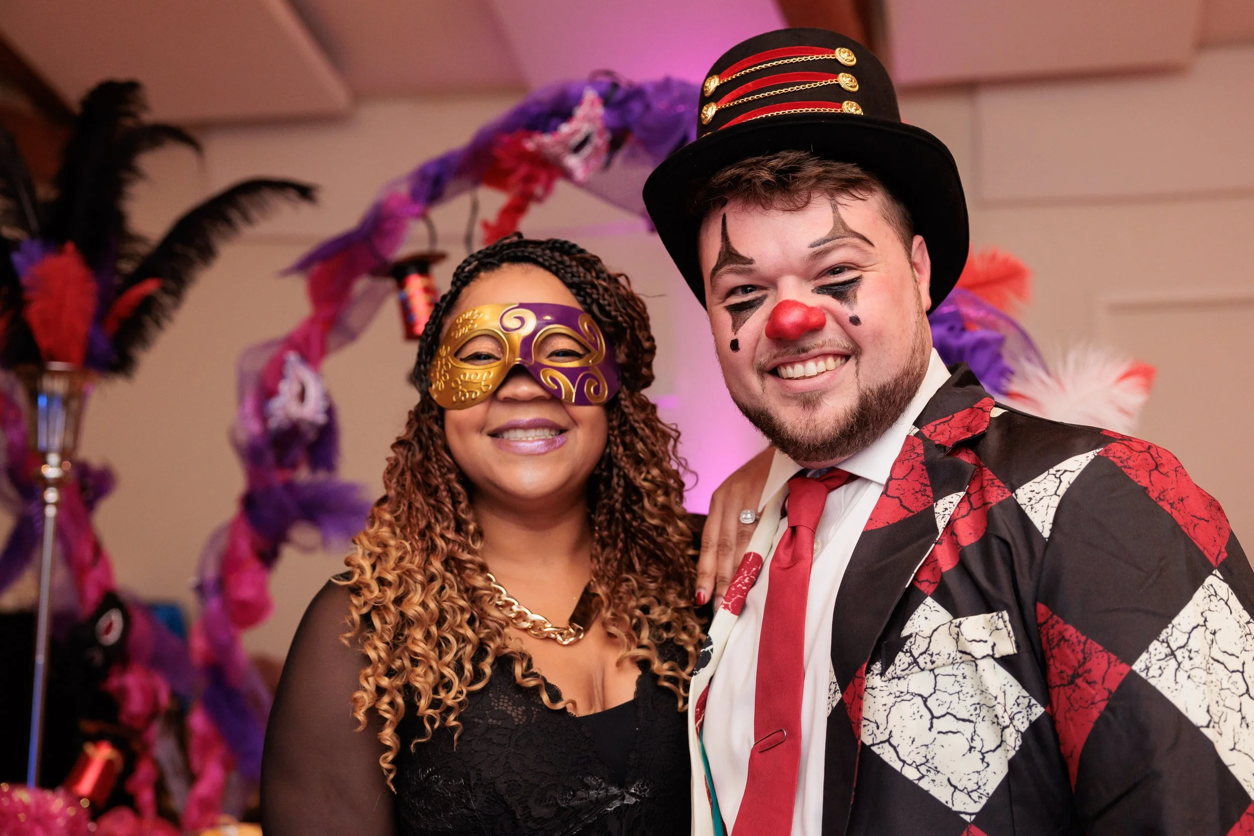 Two people at a costume party, dressed as a clown and a masked carnival performer, smiling in front of colorful decorations.