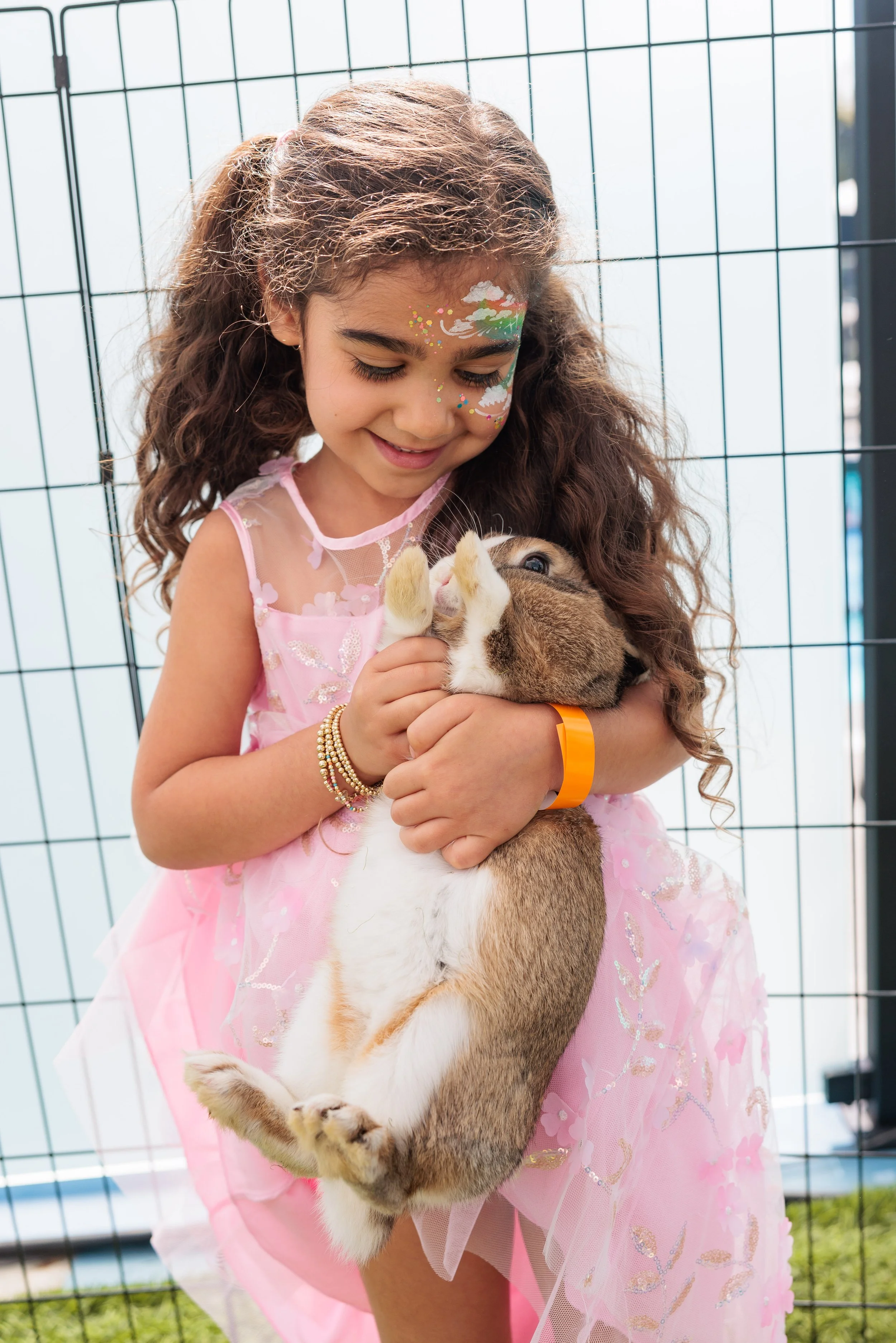 A young girl dressed in a pink dress holding a bunny rabbit, standing in front of a wire fence.