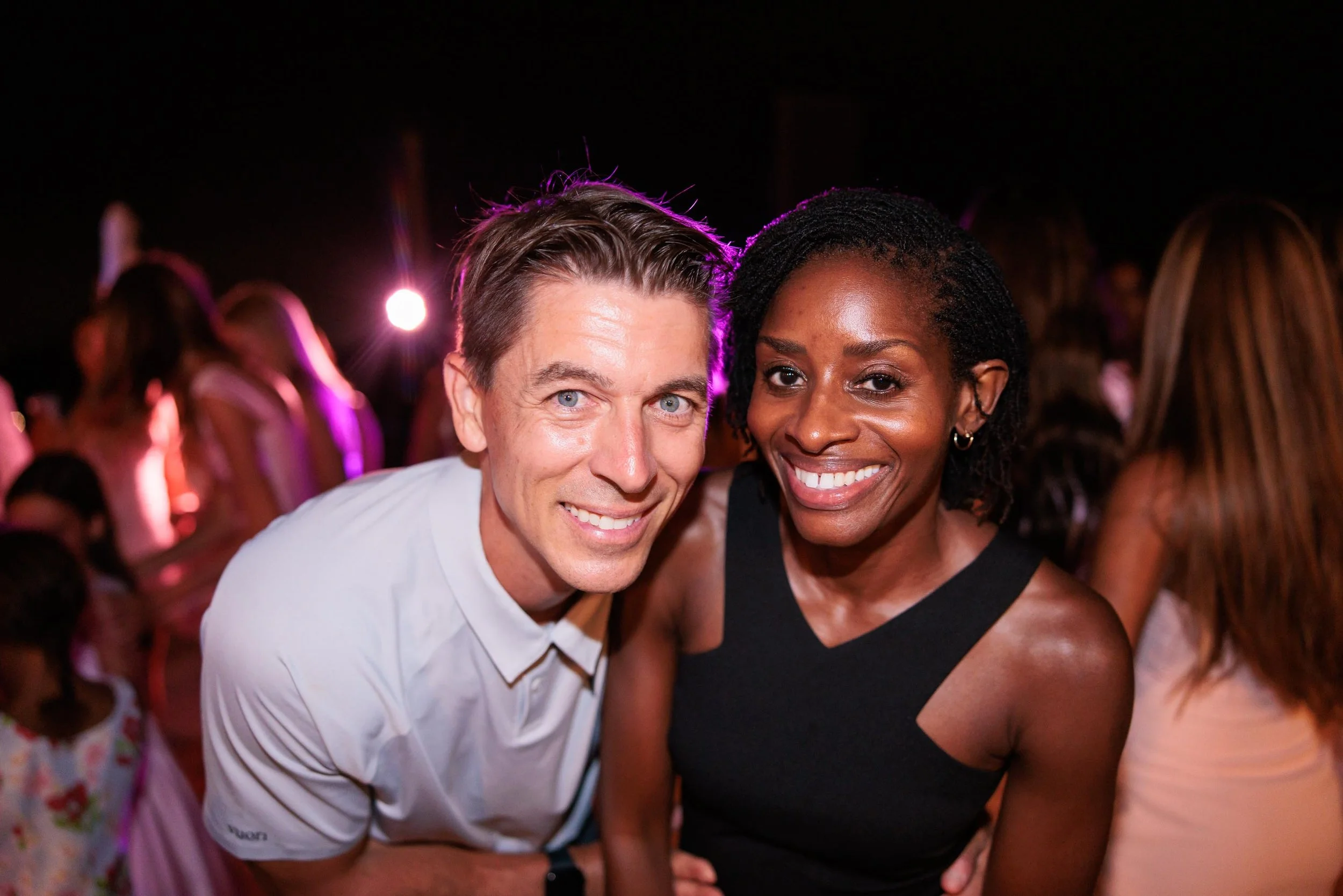 A smiling man and woman posing together at a party or social event, with colorful lights and other people in the background.