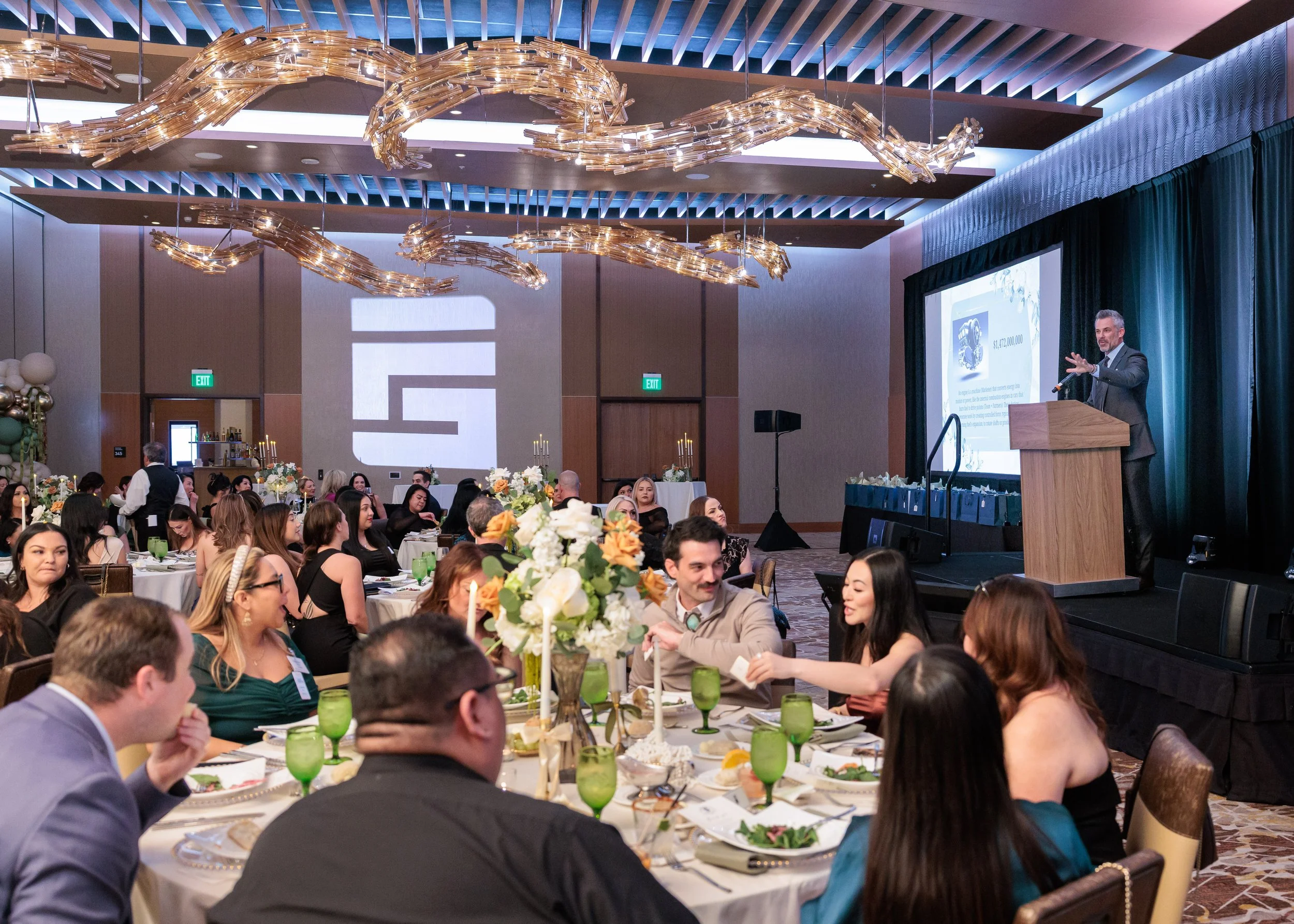 A banquet hall with round tables set for a formal event, decorated with floral centerpieces and candles. A speaker is standing at a podium on stage, presenting to the audience. The room features modern lighting fixtures and a projection screen displa