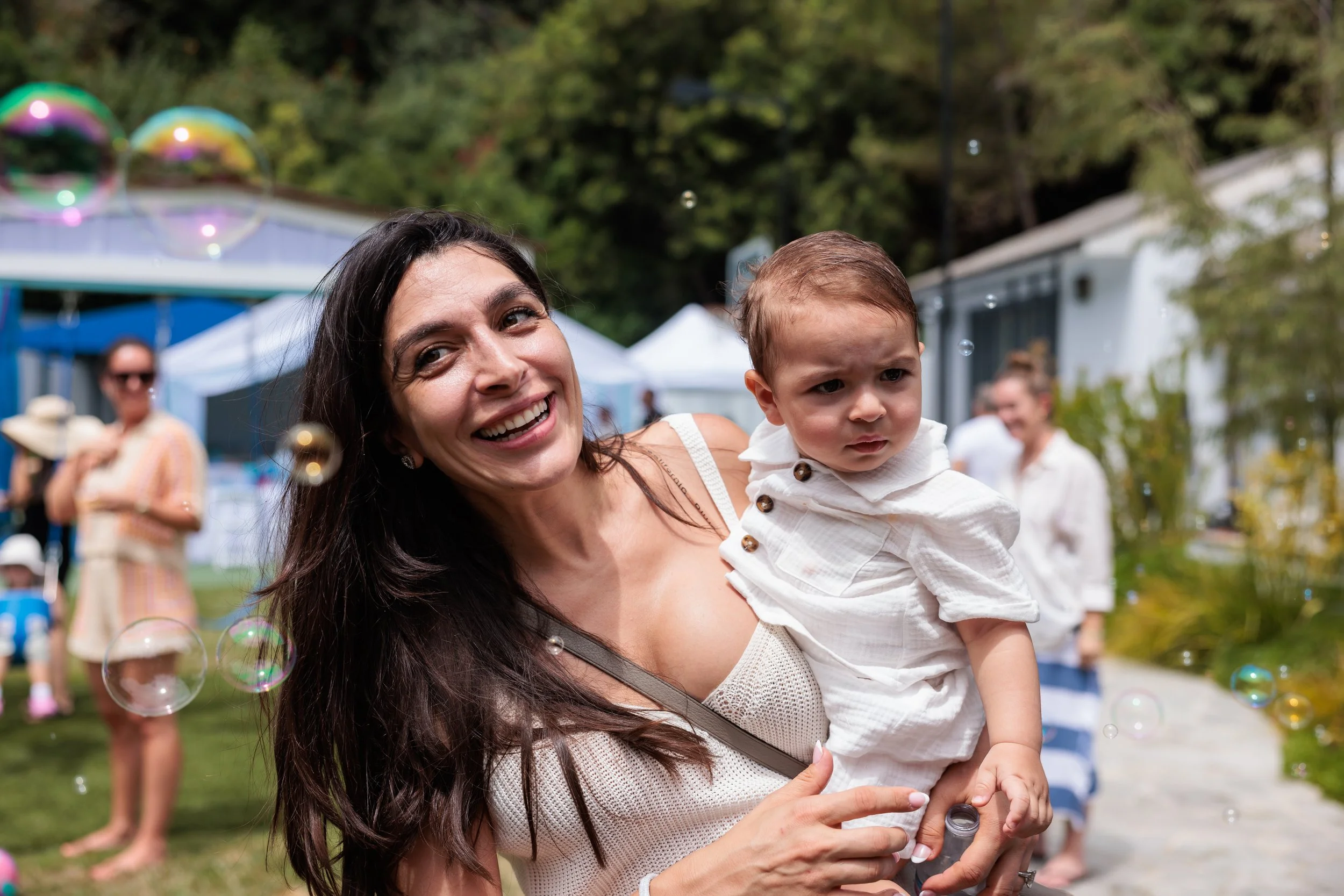 A woman smiling and holding a young boy outdoors at a party or gathering, with people and tents in the background and bubbles floating around.