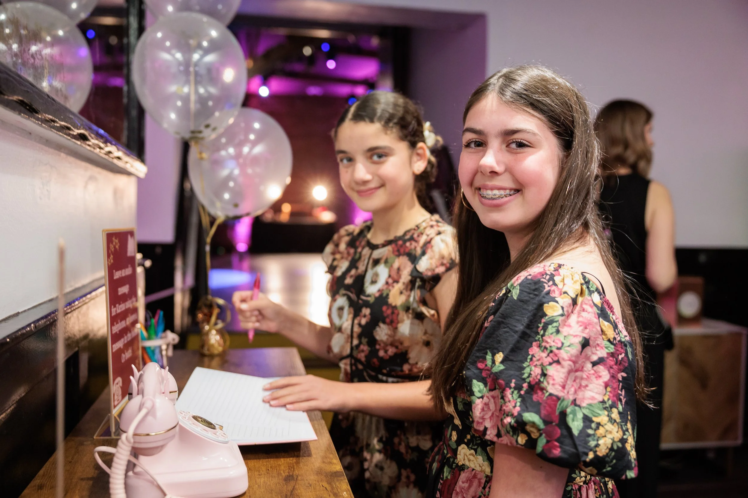 Two smiling young girls with long hair, wearing floral dresses, standing at a registration desk with a pink rotary phone and balloons in the background, at a festive event.