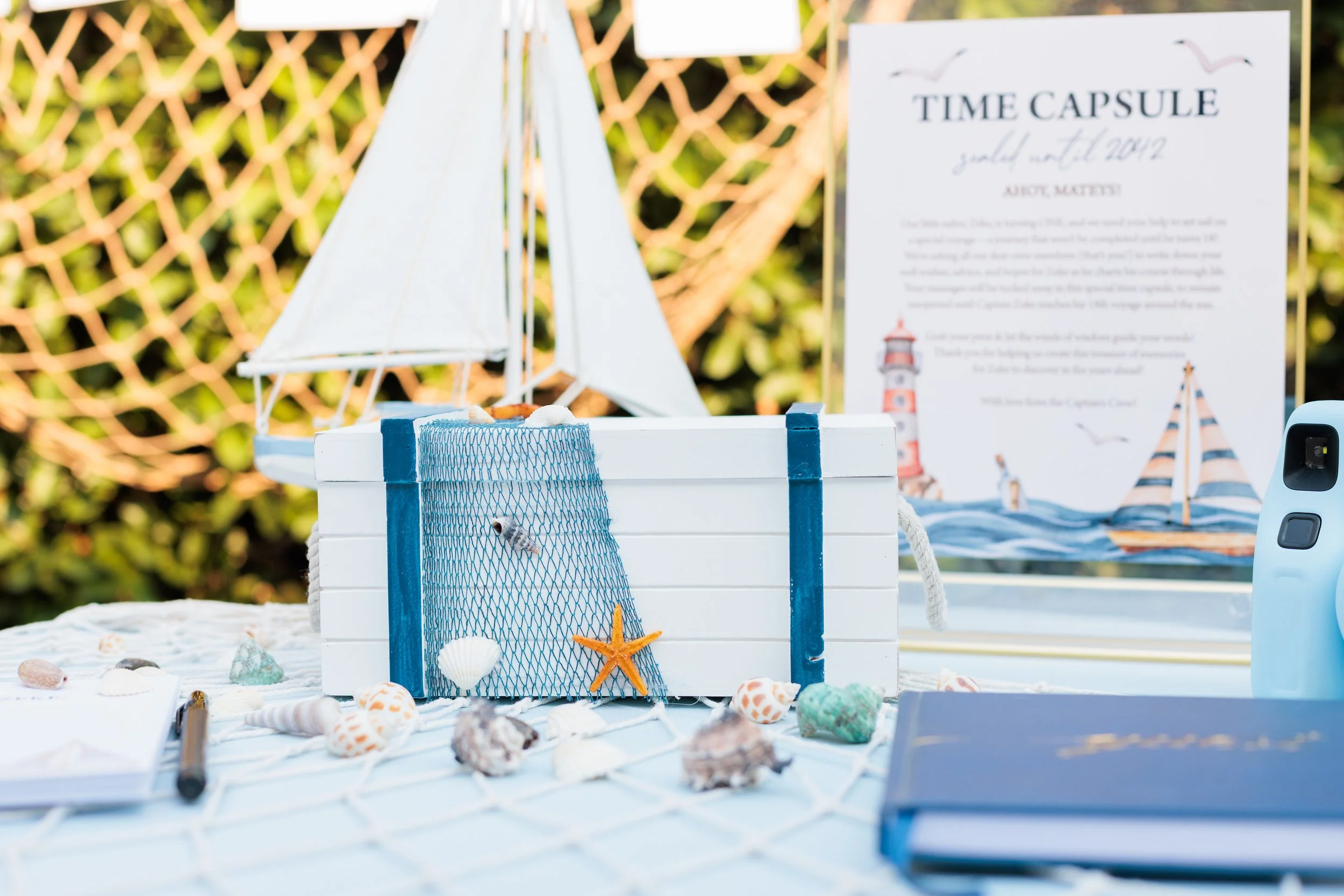 A white treasure chest decorated with a blue net and starfish, surrounded by seashells, on a nautical-themed outdoor table with a sign in the background that reads "Time Capsule July 16th, 2021."