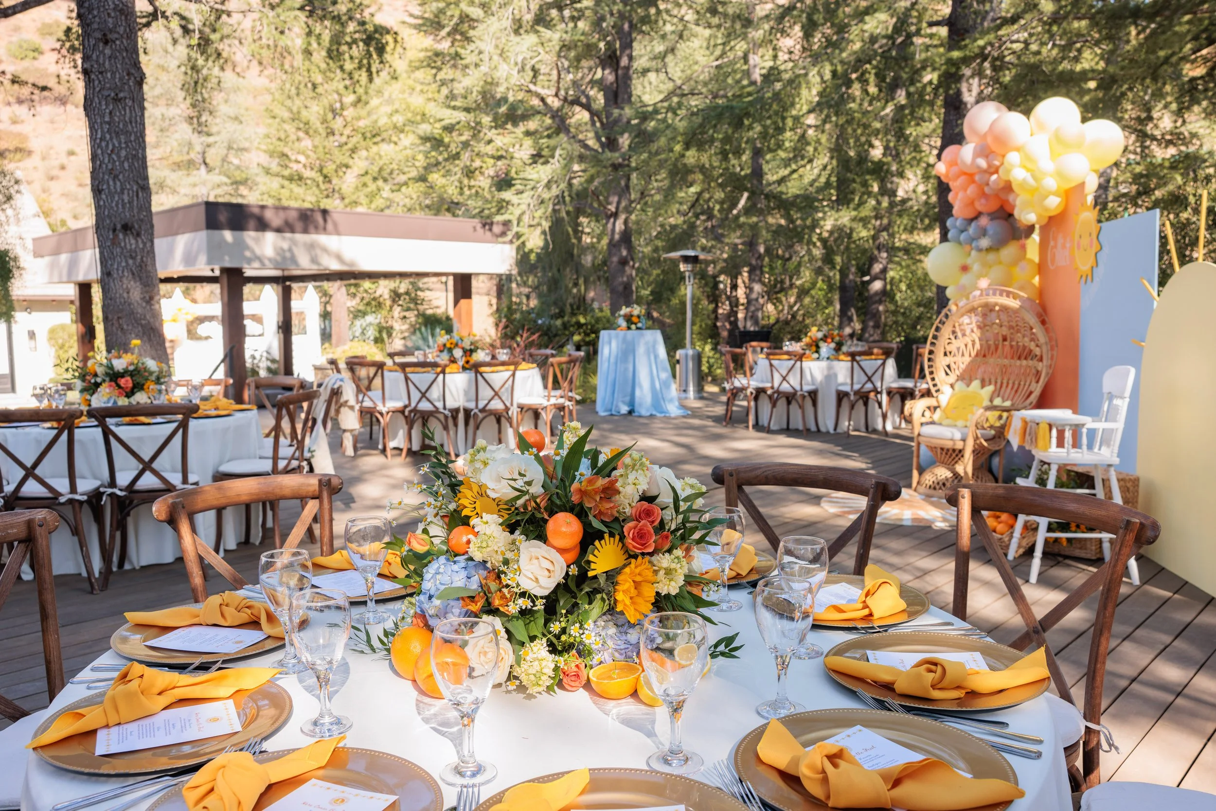 Outdoor event setup with round tables decorated with flowers and yellow napkins, surrounded by wooden chairs, and a colorful balloon and backdrop decoration in a wooded area.