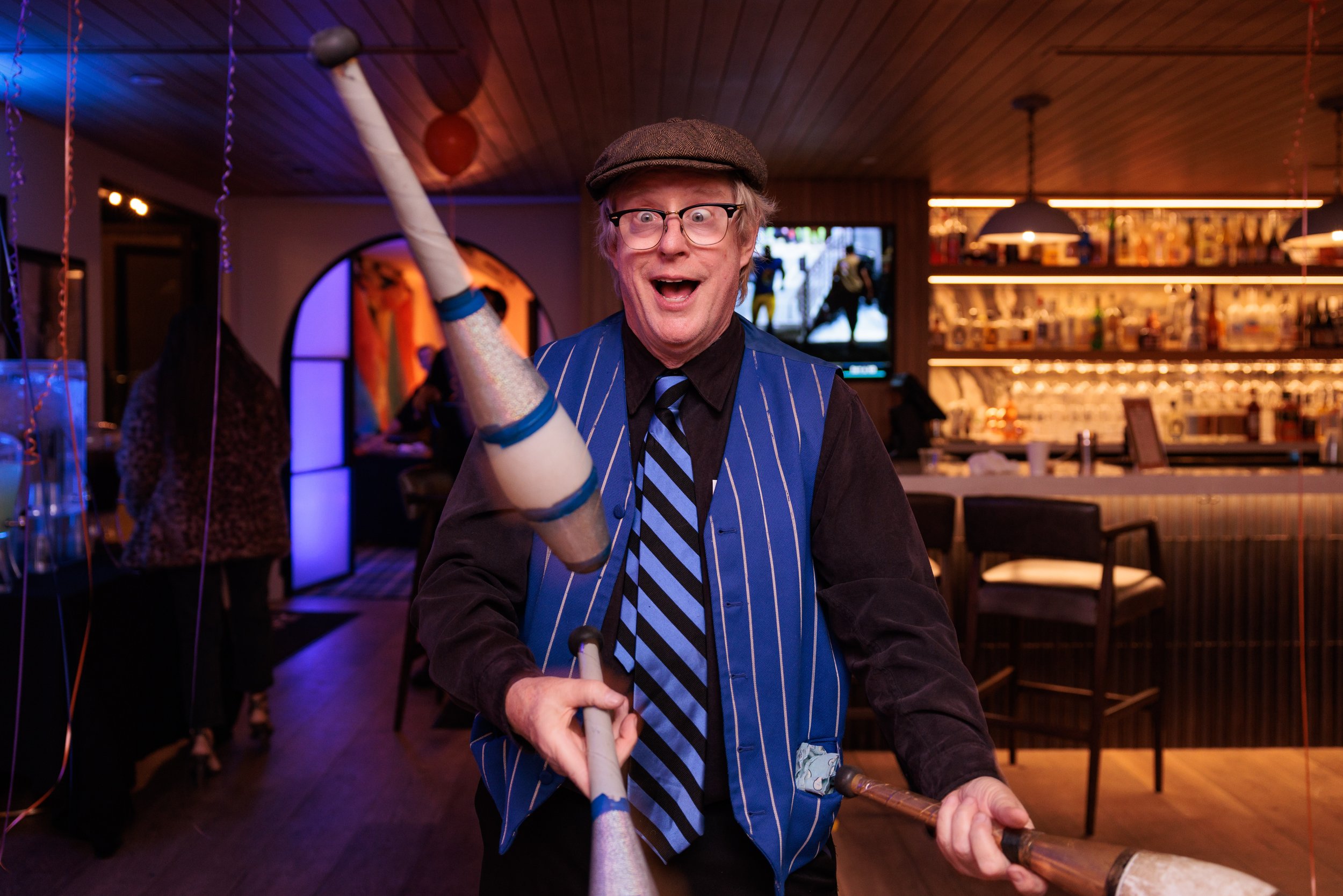 Man with glasses wearing a newsboy cap, black shirt, striped vest, and tie holding a mallet, smiling widely, at a party or celebration in a bar or restaurant with balloons and colorful lighting.