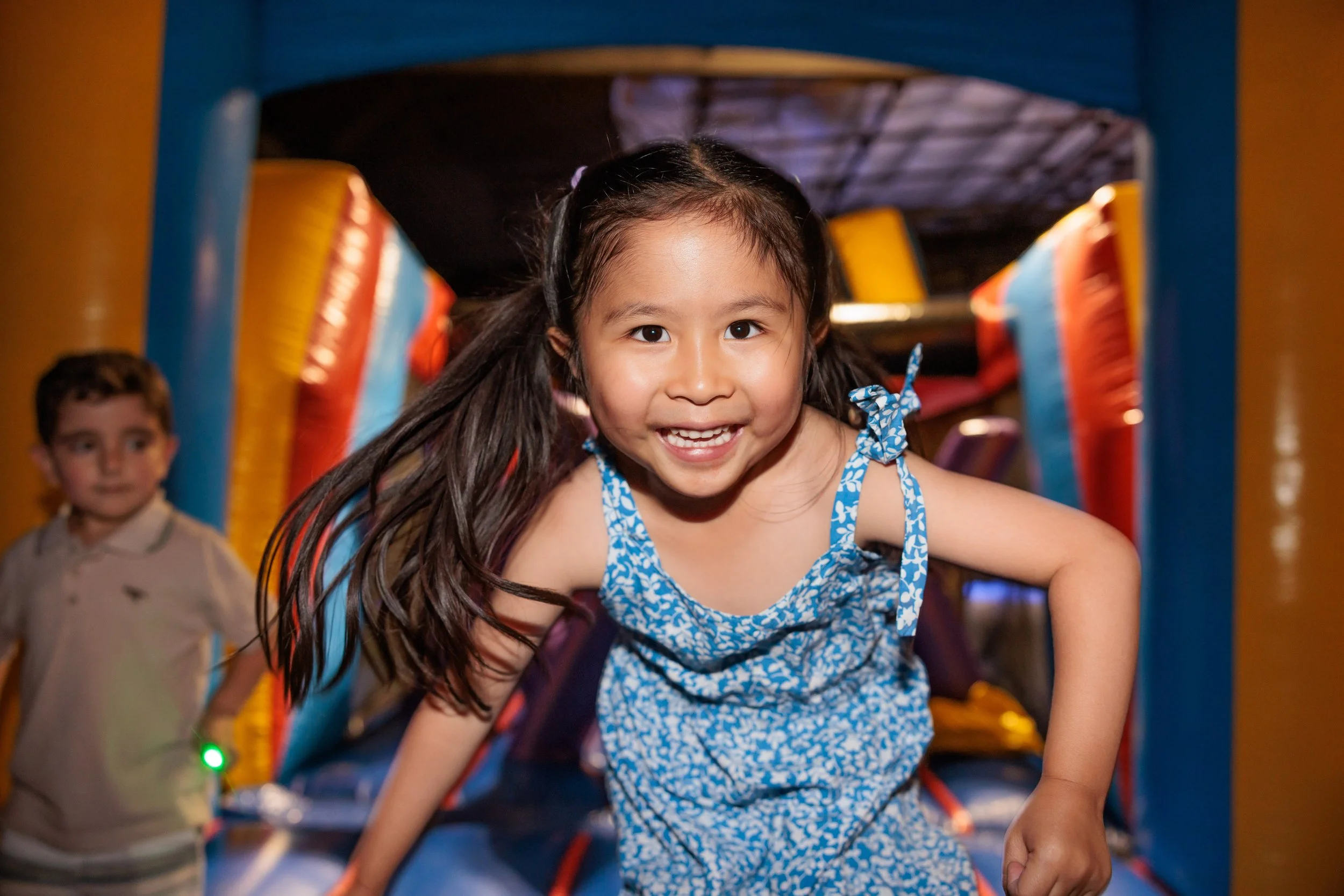 Young girl with pigtails smiling and crawling inside an inflatable bounce house with a boy watching in the background.