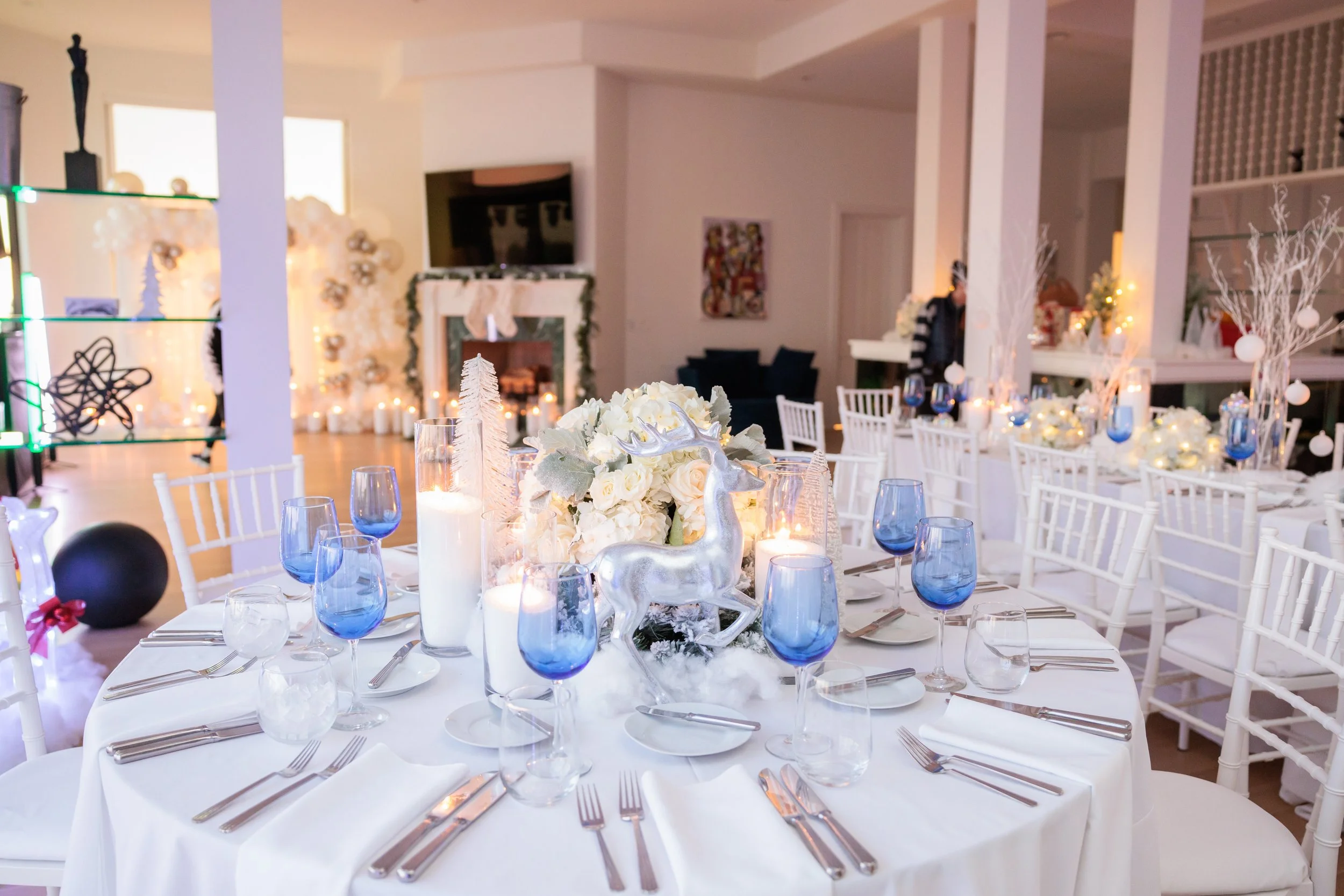 A decorated banquet table with white tablecloth, white chairs, floral centerpiece, white candles in glass holders, and blue water glasses, set for a holiday or winter celebration.