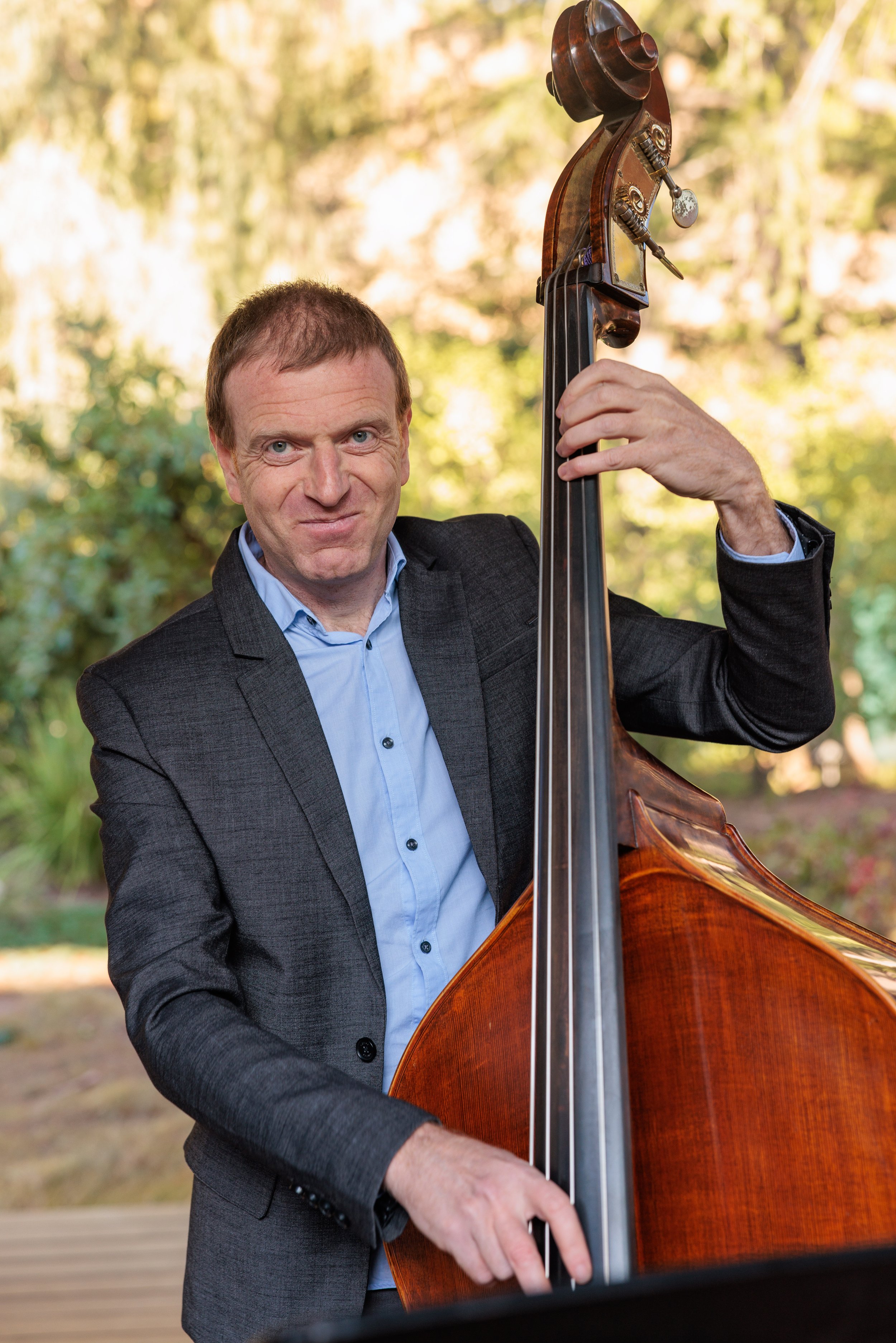 A man in a dark gray suit and light blue shirt playing a double bass outdoors, with trees and sunlight in the background.