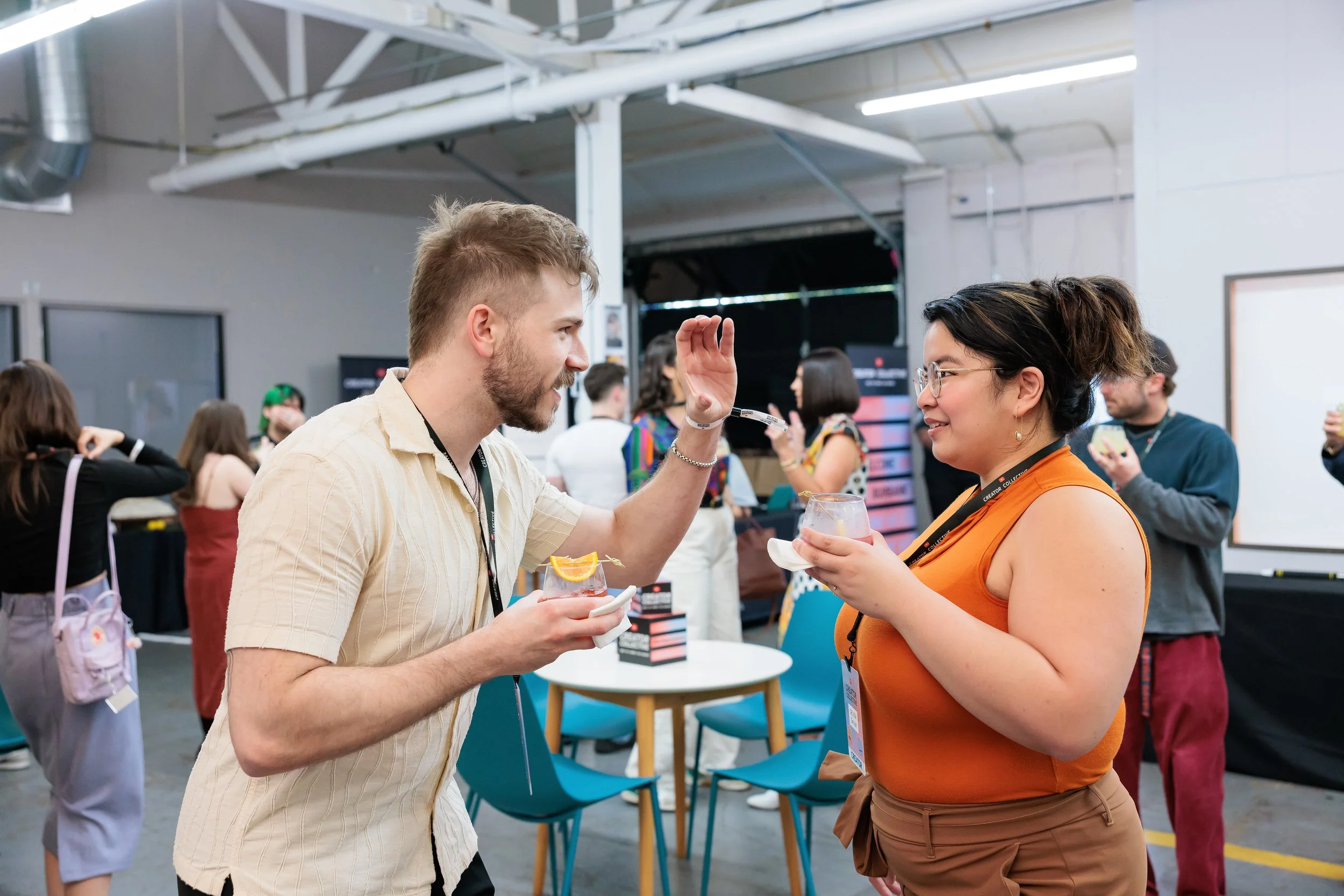 A man and woman talking at a social event, holding drinks, with multiple people in the background in an indoor venue.