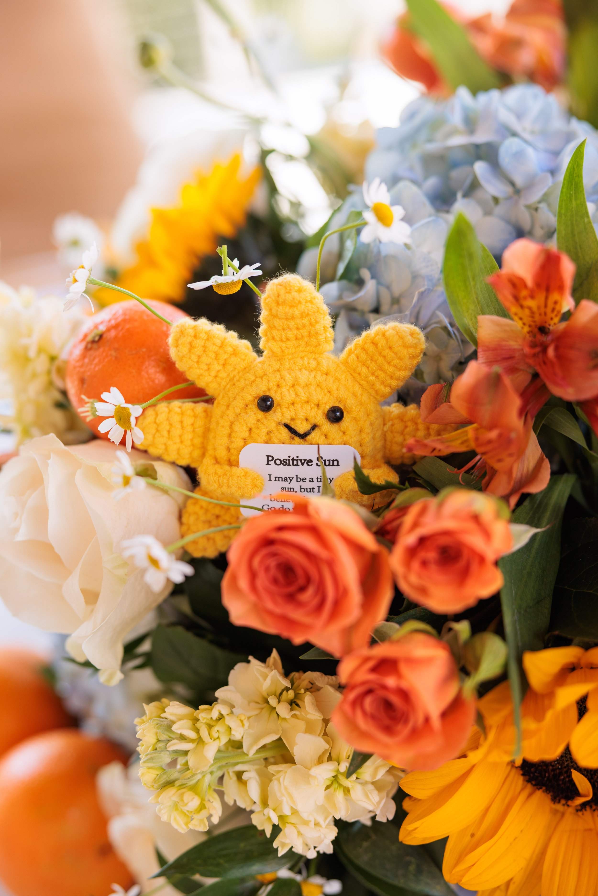 A colorful bouquet of flowers including sunflowers, roses, hydrangeas, and daisies, with a yellow knitted Pikachu plush toy in the center. The plush has black eyes, a stitched smile, and a tag that reads "Positive Sun".