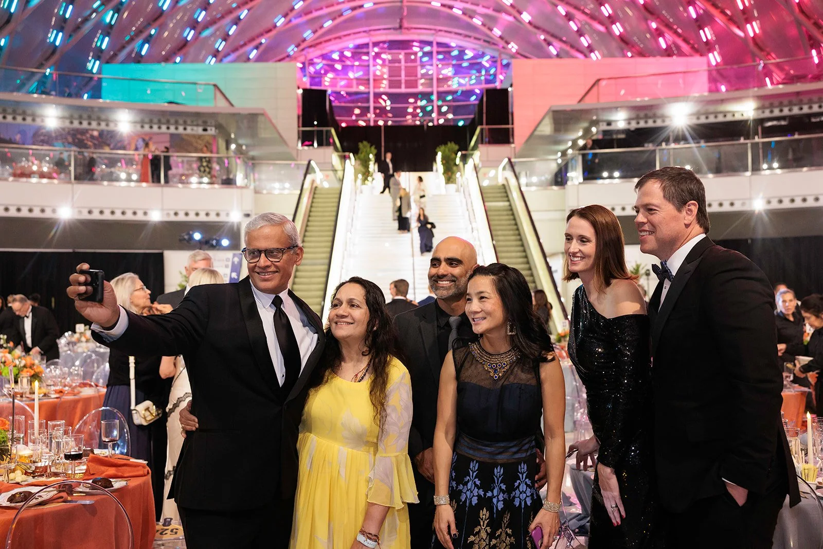 Group of friends in formal attire taking a selfie at a fancy event or banquet, with a grand staircase and colorful lit ceiling in the background.