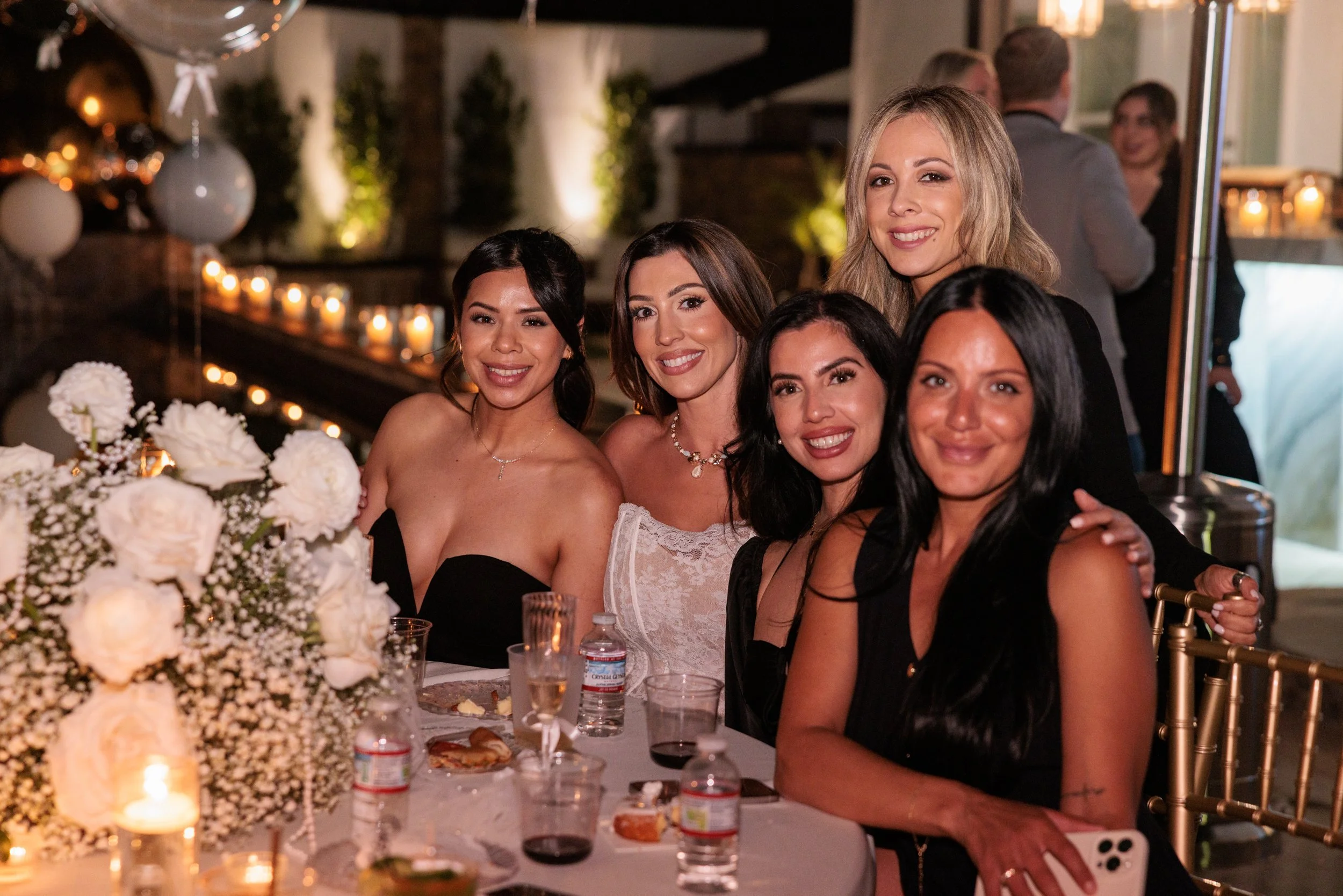 Five women in elegant attire smiling at a decorated evening party with candles, flowers, and balloons.