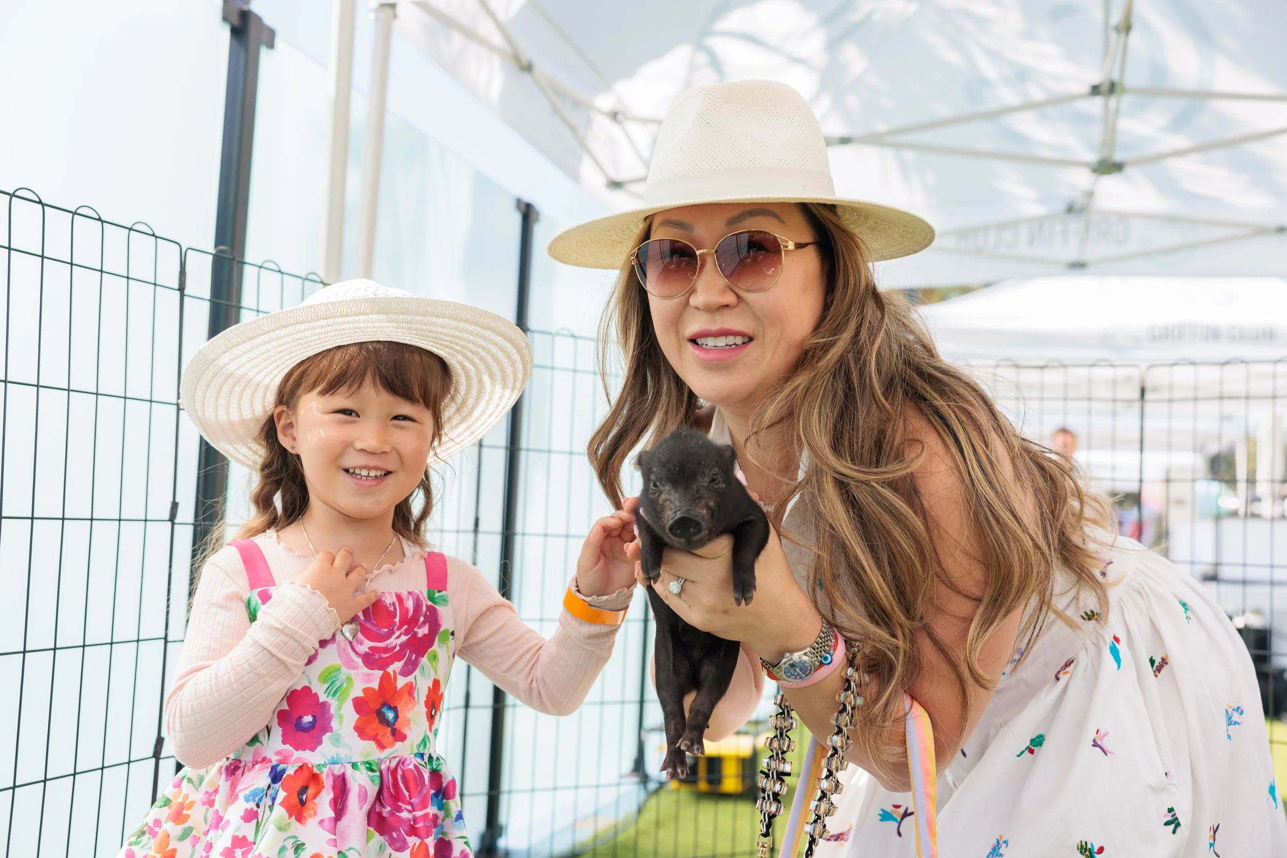 A woman and a young girl at an outdoor event holding a small black puppy. They are both wearing wide-brimmed hats and smiling, with a wire fence and white canopy tents in the background.