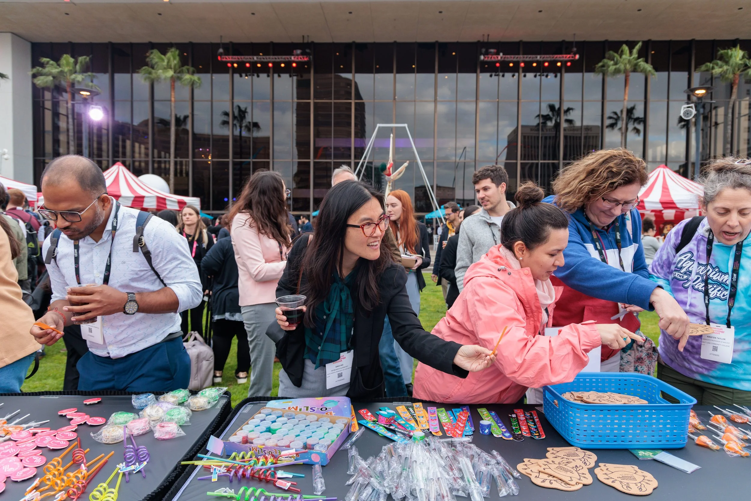 People gathered at an outdoor event table filled with colorful party favors, candy, and toys. Four women at the front of the table are engaging with the items, one holding a cup of drink and others reaching for toys and candies. In the background, mo