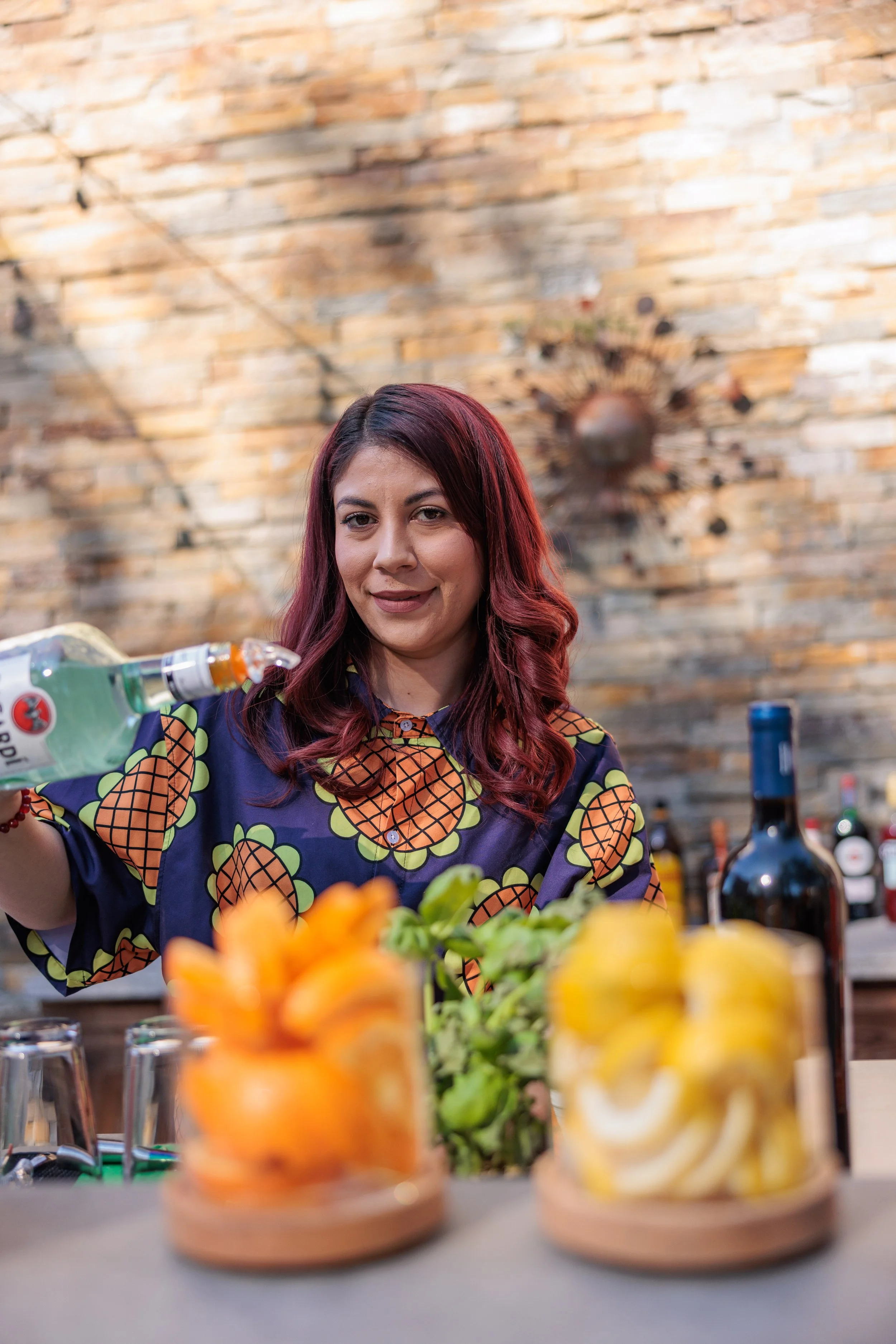 A woman with red hair and a pineapple-patterned shirt pours a drink at a bar or cafe with a brick wall background. There are glasses and jars of fruit on the counter.