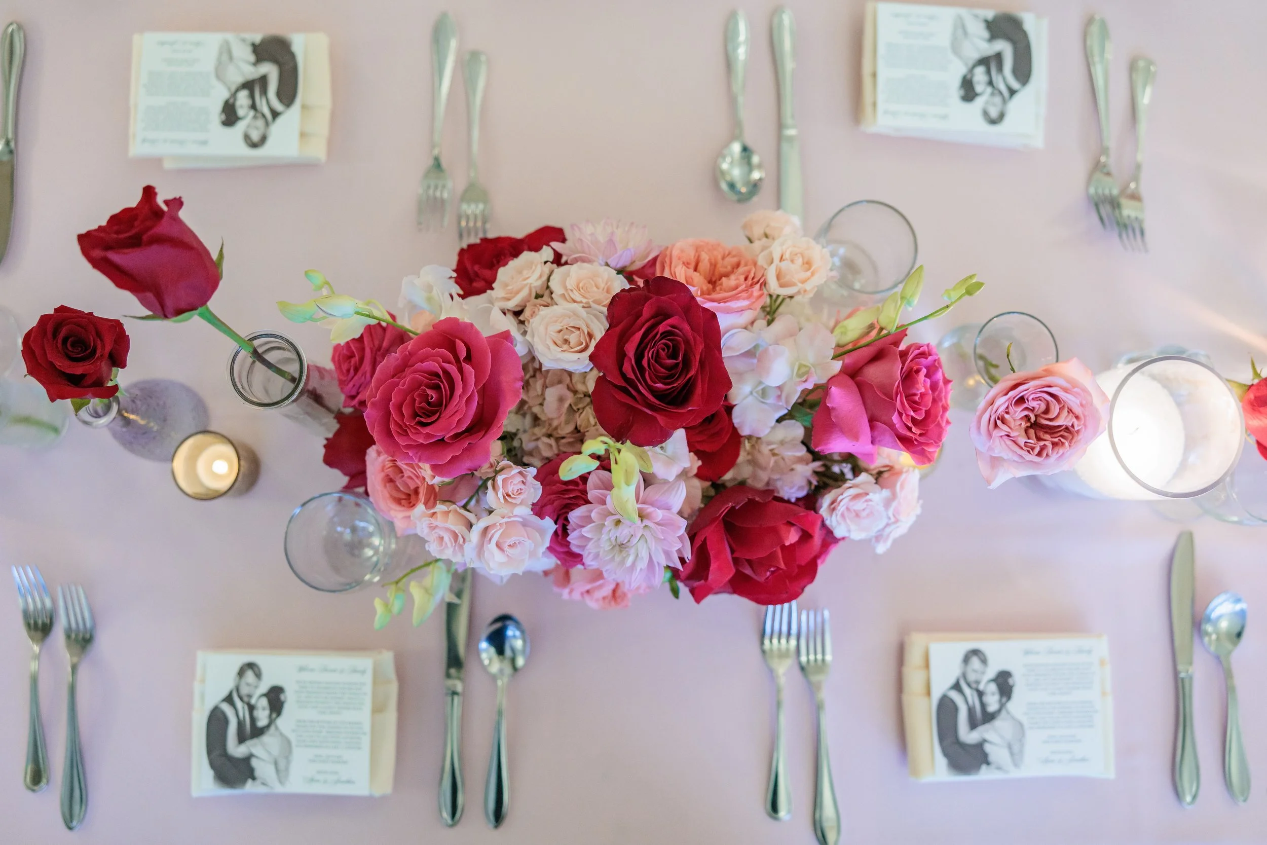 A dining table with a centerpiece of various pink, red, and white flowers, surrounded by utensils and candleholders, with printed menus or place cards.