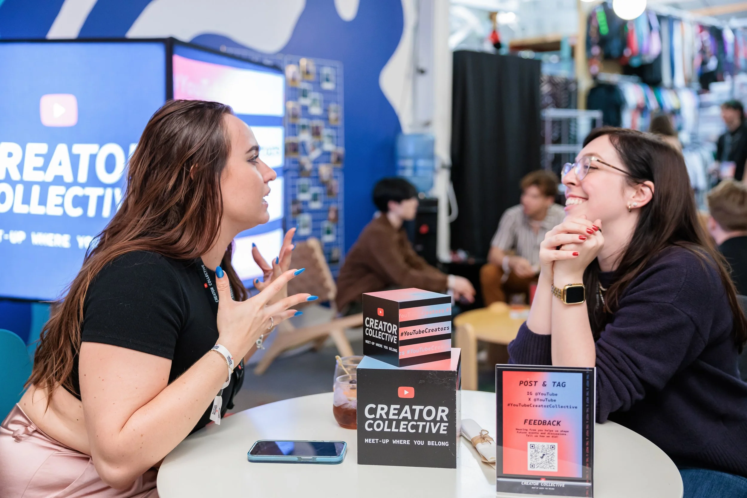 Two women engaging in conversation at a table during a creator community event, with a large YouTube Creator Collective sign and promotional materials on the table.