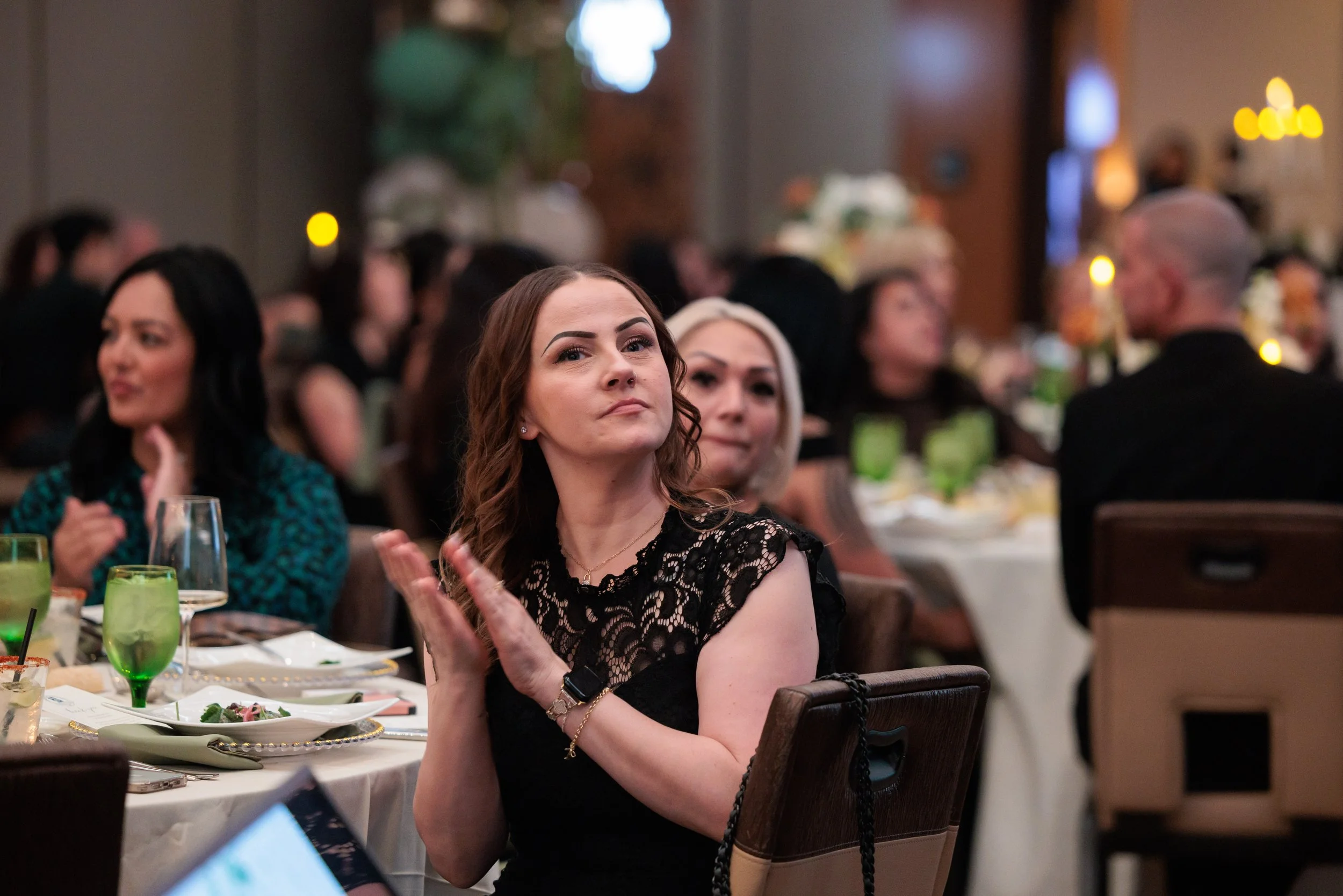 A woman with brown hair in a black lace dress is clapping at a formal event or dinner, surrounded by other guests seated at tables with food and drinks, in a decorated banquet hall.
