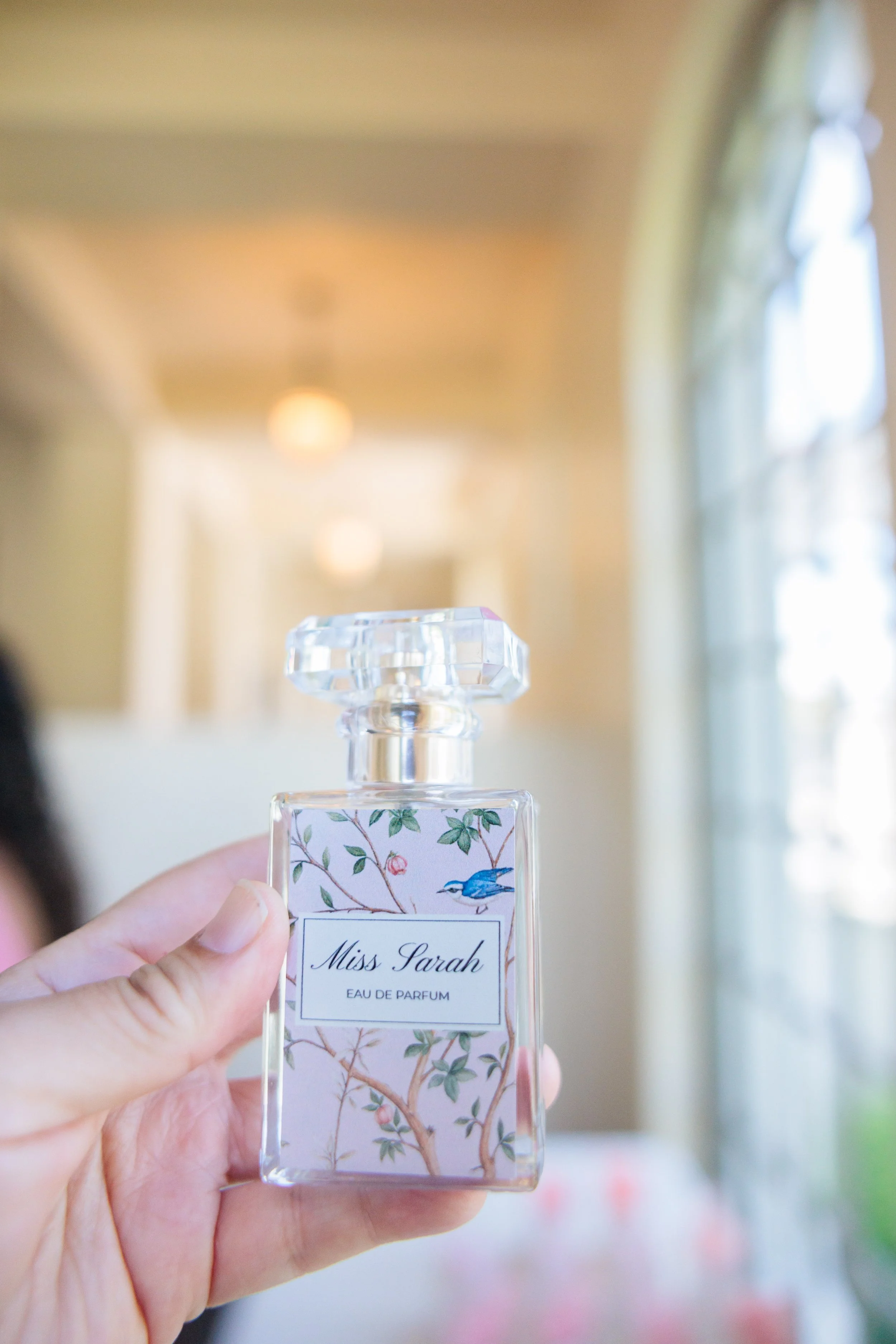 Close-up of a hand holding a bottle of Miss Sarah Eau de Parfum with floral and bird design, blurred background with windows and warm lighting.