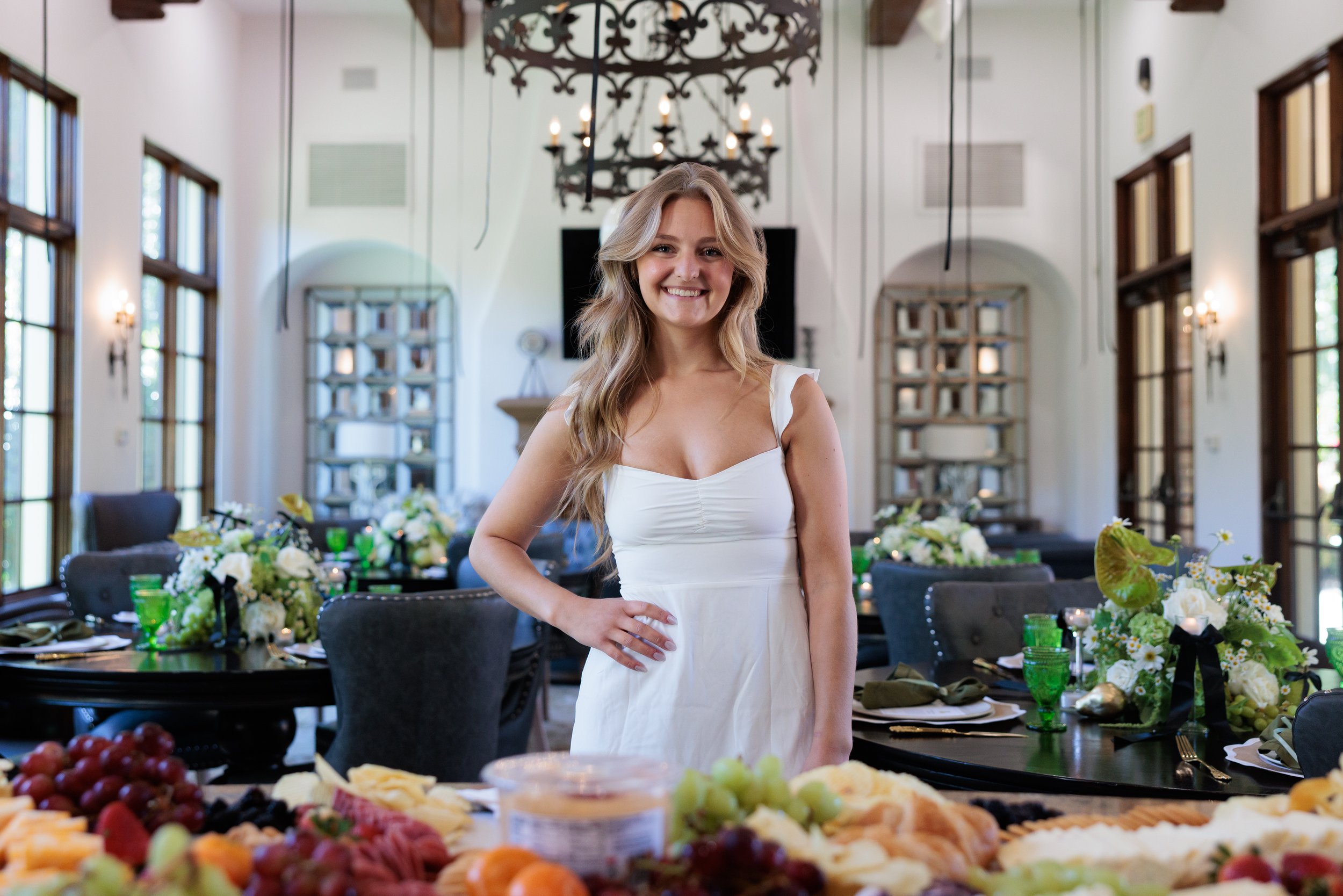 A young woman in a white dress smiling at a feast table with fruit and cheese, in a decorated dining room.